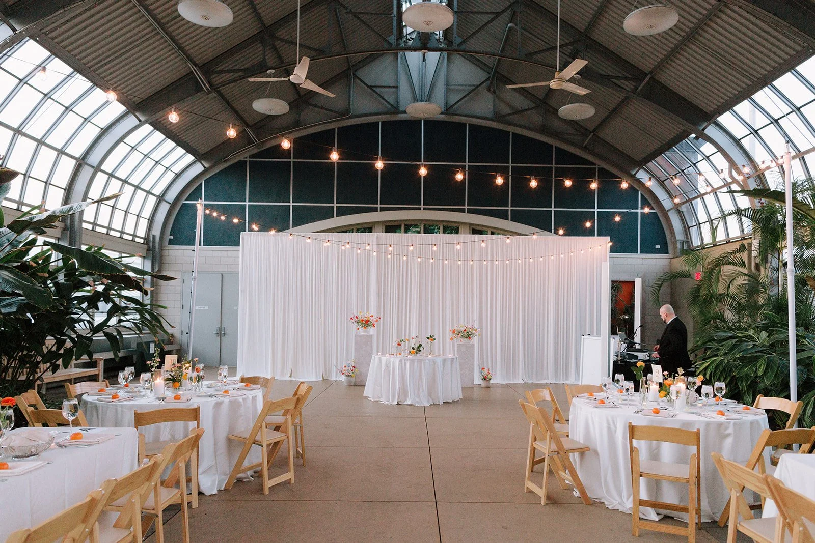 Indoor event space with round tables decorated with white tablecloths and floral centerpieces. The space has a high, arched glass ceiling with string lights hanging across. A white curtain backdrop with floral arrangements is at the front. There are 