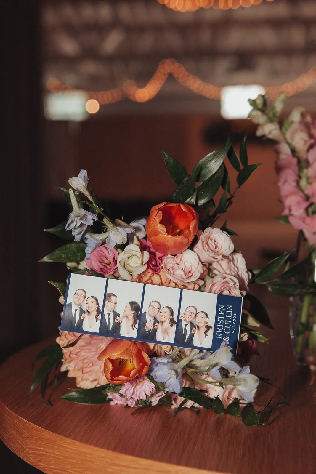 Flower arrangement with a photo booth strip and text indicating a wedding between Kristen and Cullen on April 5, 2024, with a background of string lights.