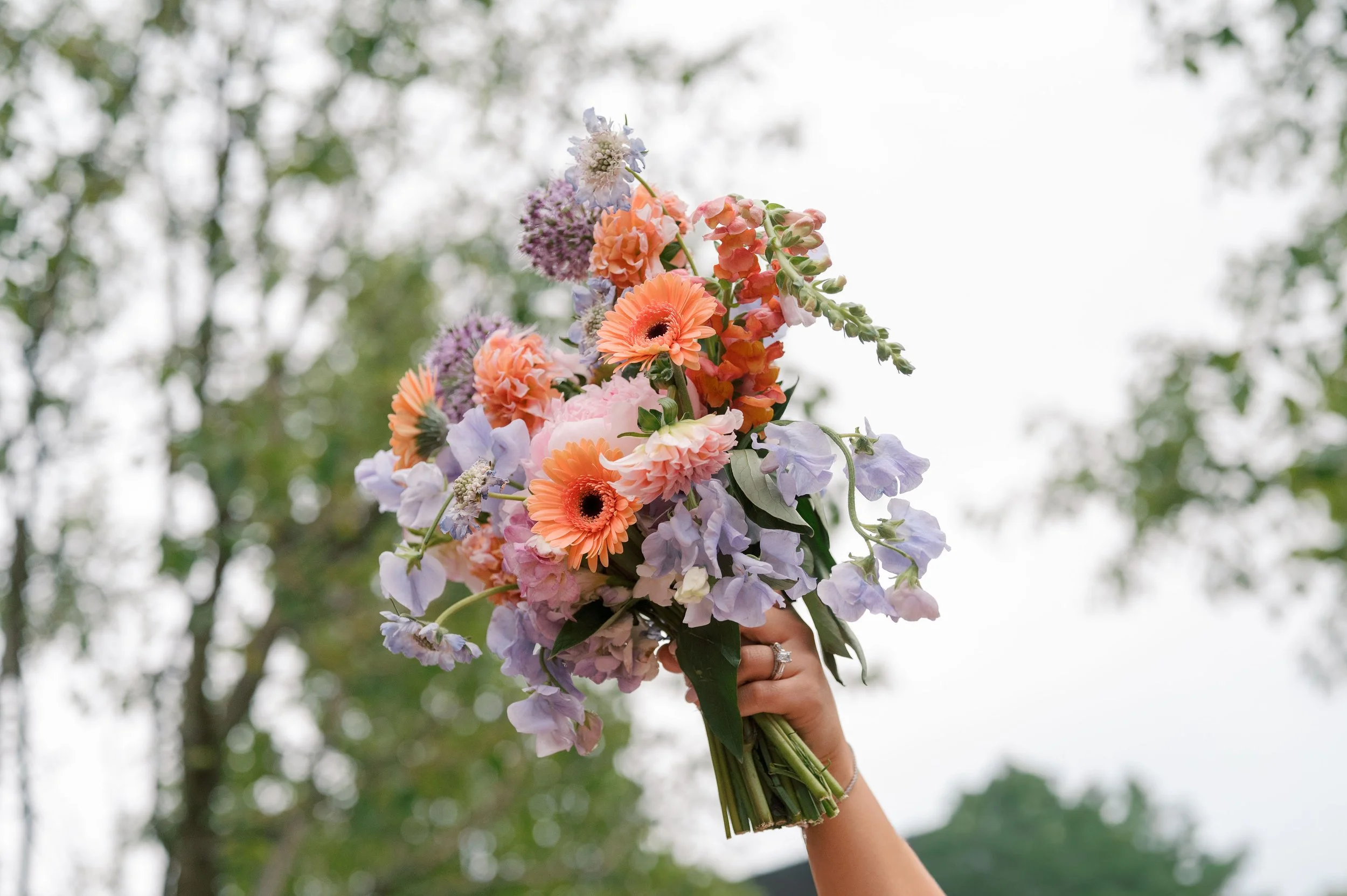 A person holds a bouquet of flowers with peach, pink, purple, and white blossoms against a background of blurred green trees.