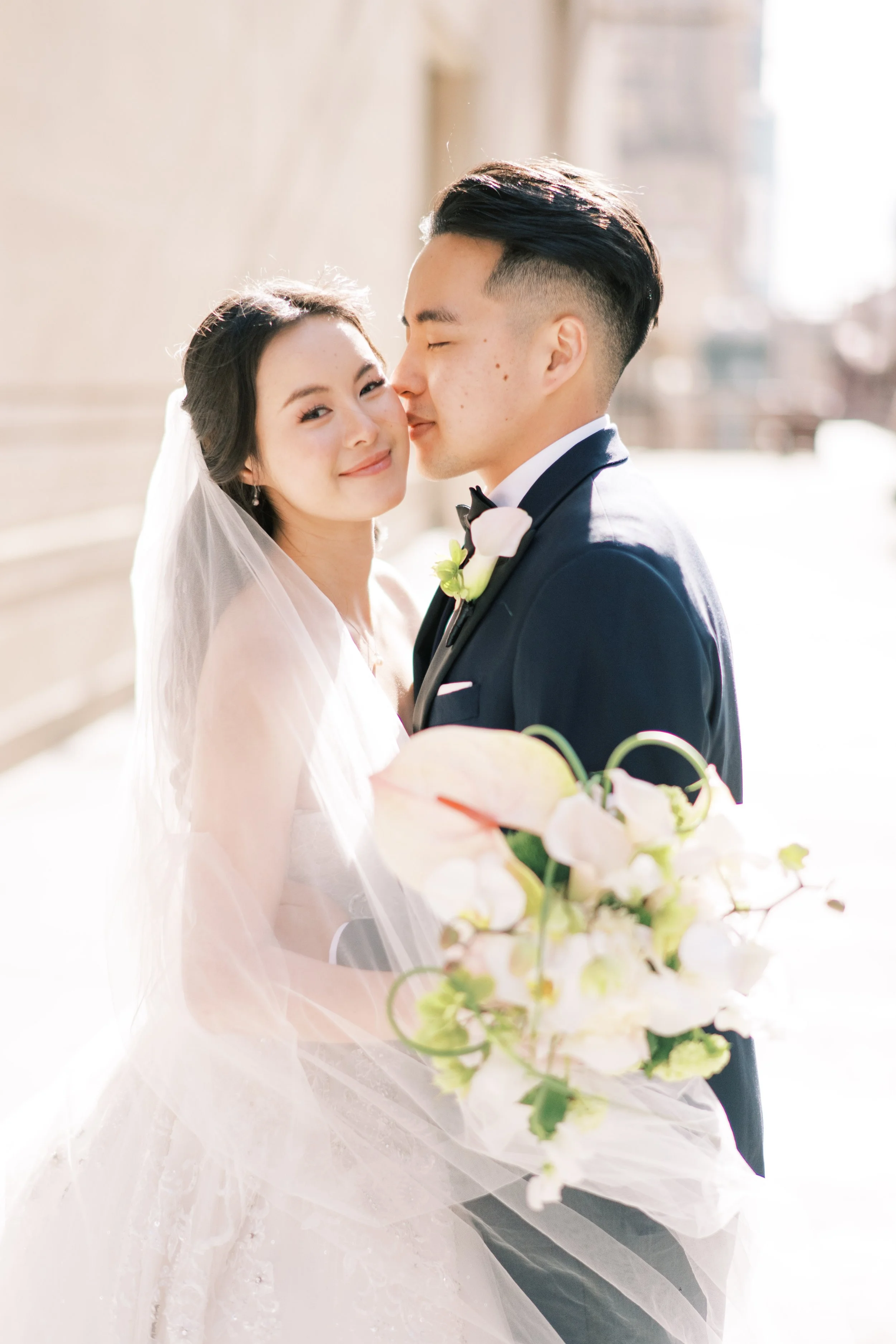 A bride and groom on their wedding day, standing outdoors with sunlight shining behind them. The bride is smiling and looking at the camera, wearing a white dress and veil. The groom is kissing the bride's cheek, dressed in a black tuxedo with a bout