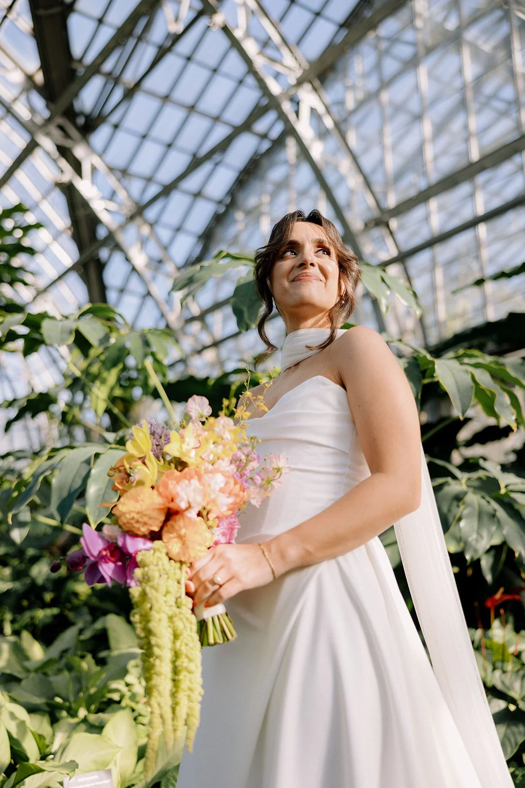 A woman in a white dress holding a bouquet of colorful flowers inside a greenhouse, looking off into the distance with a smile.