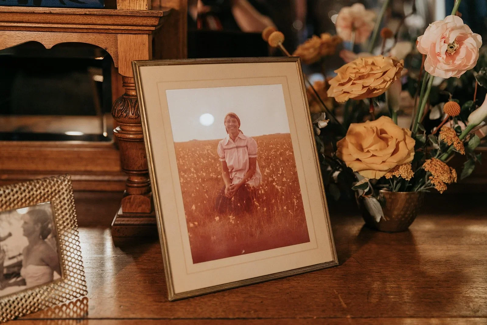A framed photograph of a woman sitting in a field, with the sun setting behind her, placed on a wooden table alongside a small black and white photo in a woven frame and a colorful flower arrangement in a bronze vase.