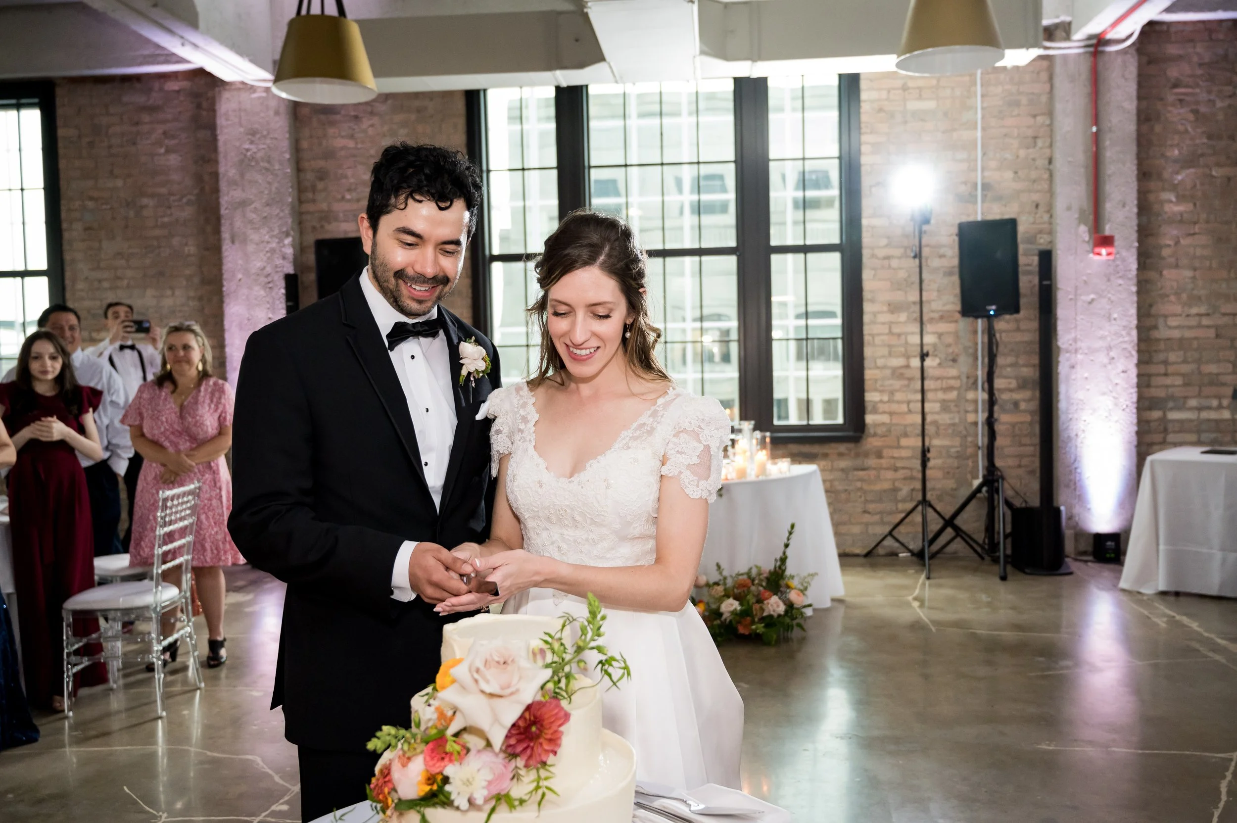 A newlywed couple cutting a wedding cake together during a wedding reception in an industrial-style venue with large windows and exposed brick walls. The groom is wearing a black tuxedo with a bow tie, and the bride is in a white lace wedding dress. 