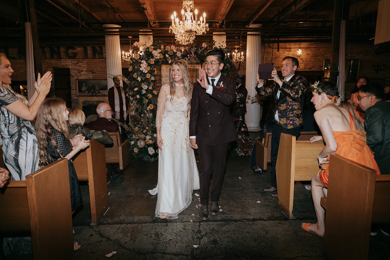 A wedding ceremony with the bride and groom walking down the aisle, surrounded by guests who are clapping and taking photos. The setting is decorated with flowers and chandeliers.