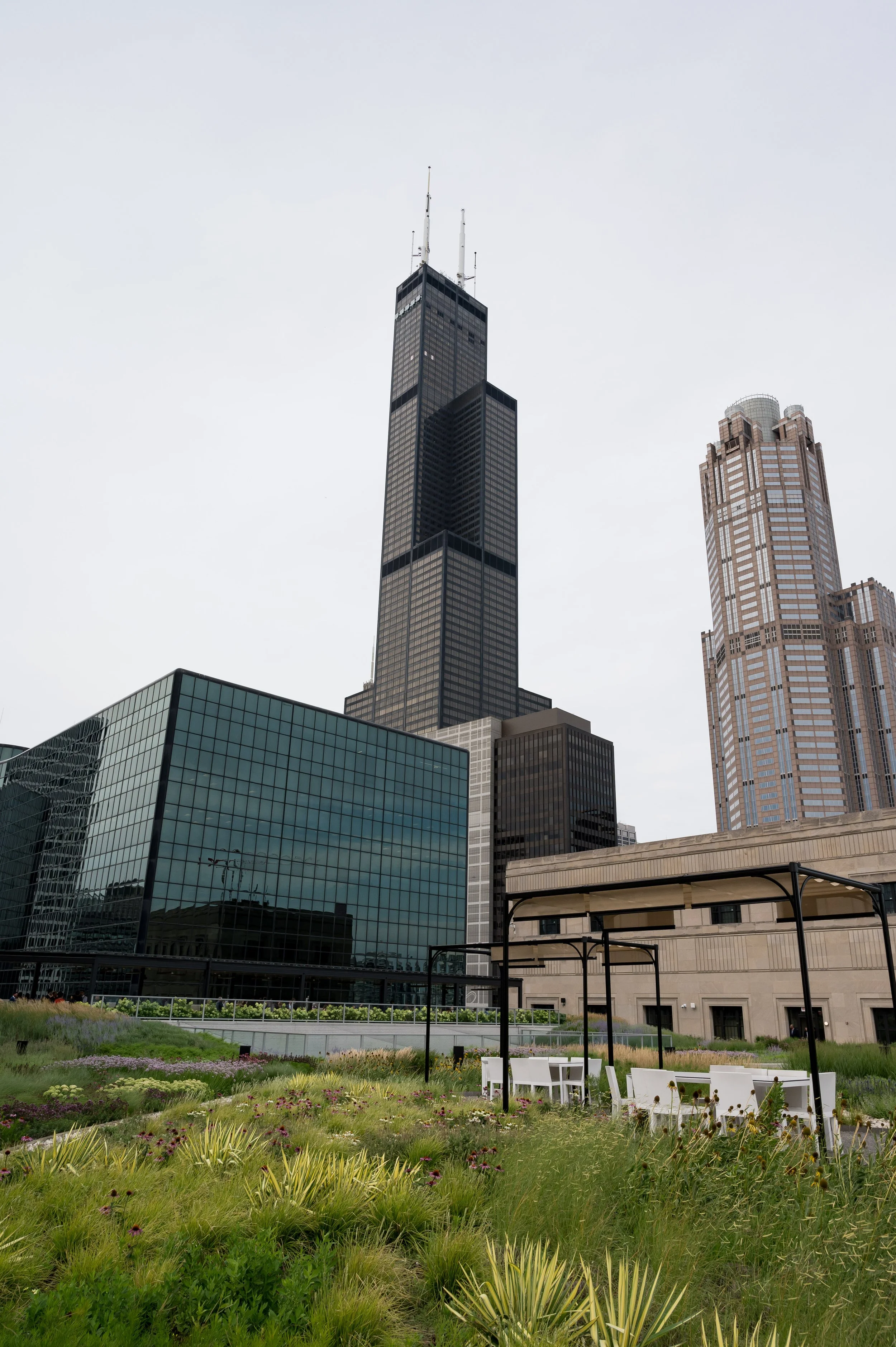 A cityscape featuring a tall black skyscraper, adjacent to a beige high-rise, with a glass building and a landscaped garden in the foreground.