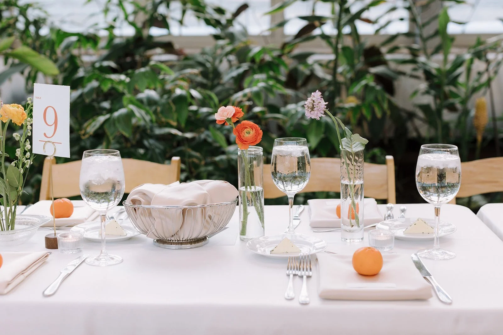 Elegant table setting with glasses of water, flowers in vases, a basket of cloth napkins, and a table number sign, with a lush green backdrop.