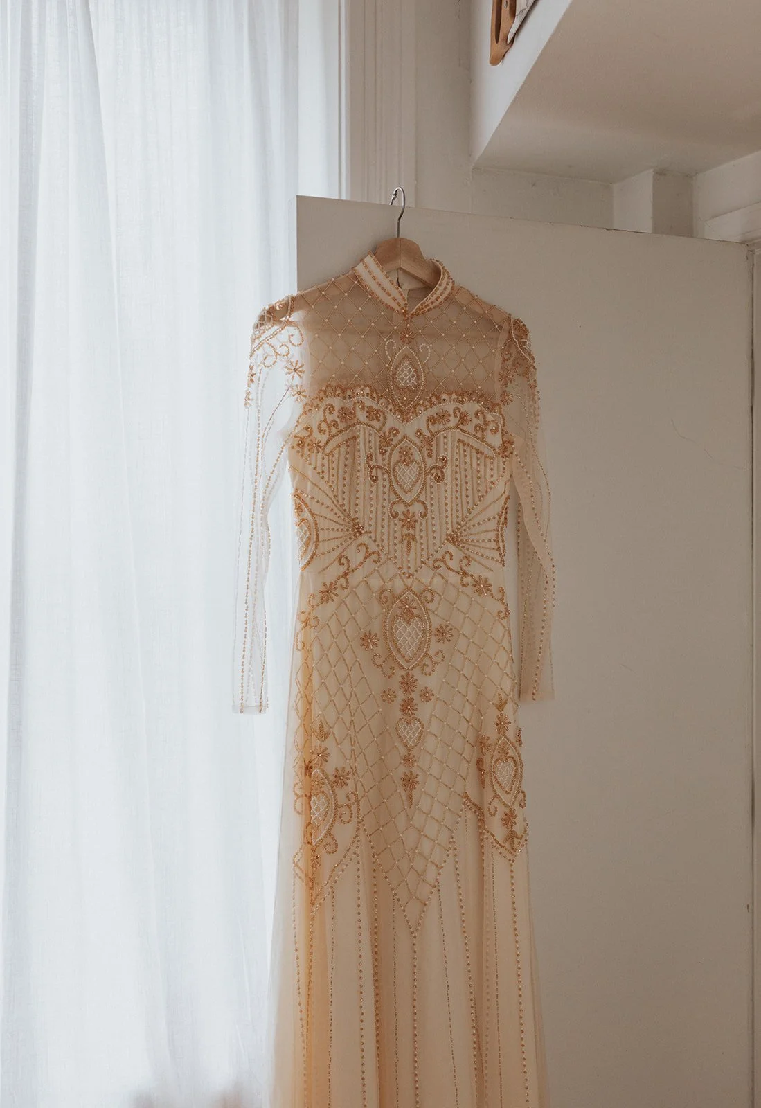 An ornate, cream-colored dress with gold embroidery hanging on a wooden hanger in front of sheer white curtains.