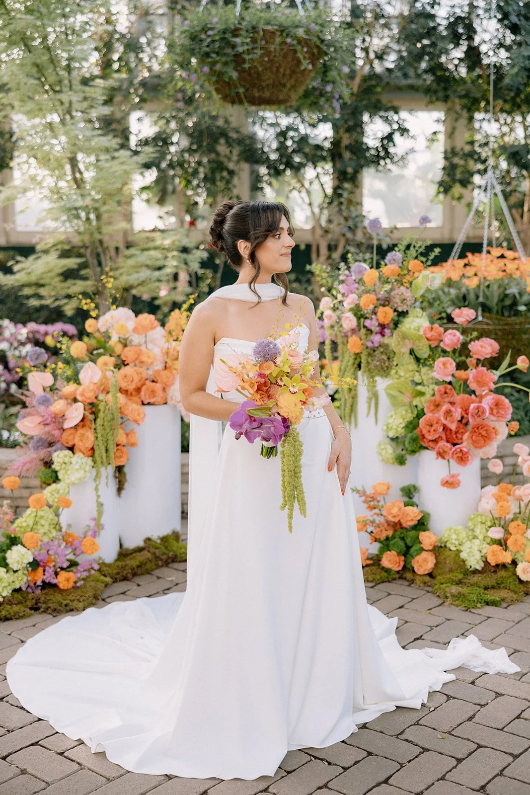 A woman in a white dress holding a colorful bouquet, standing in front of a floral wedding arch with pink, orange, purple, and green flowers.