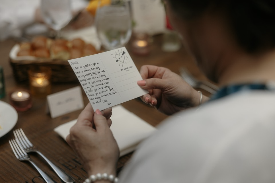 A person sitting at a table holding a small handwritten note card with a drawing of a sun. The table has plates, utensils, and glasses, and a basket of bread or pastries in the background.