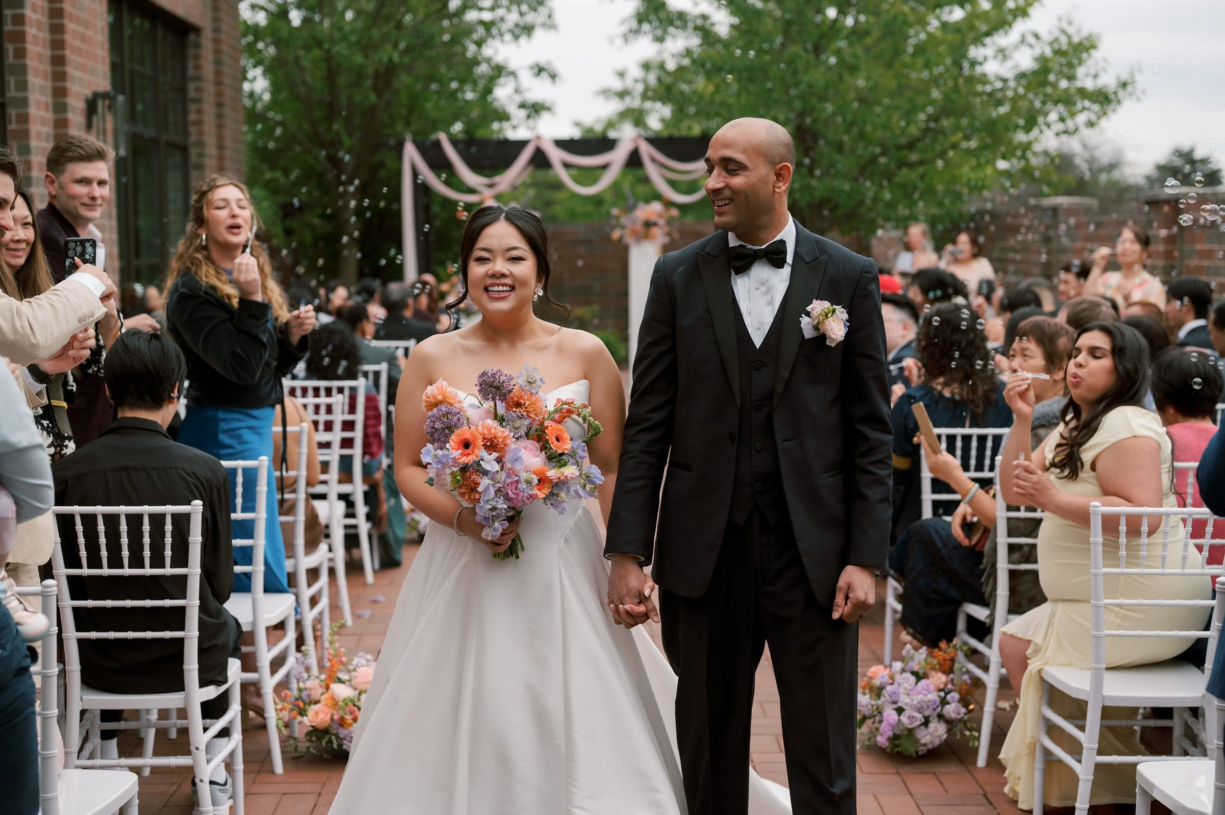 A bride and groom holding hands and smiling during their wedding ceremony outdoors, surrounded by seated guests and floral decorations.