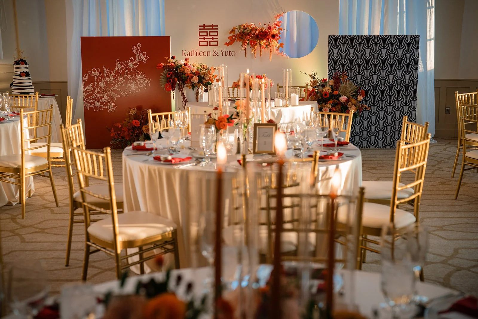 Elegant wedding reception setup with round tables, gold chairs, floral centerpieces, and a decorated backdrop with the names Kathleen and Yuto.