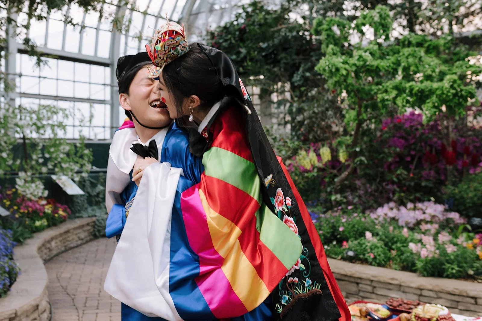 Two people in colorful traditional Korean hanbok embracing and smiling inside a greenhouse with blooming flowers.