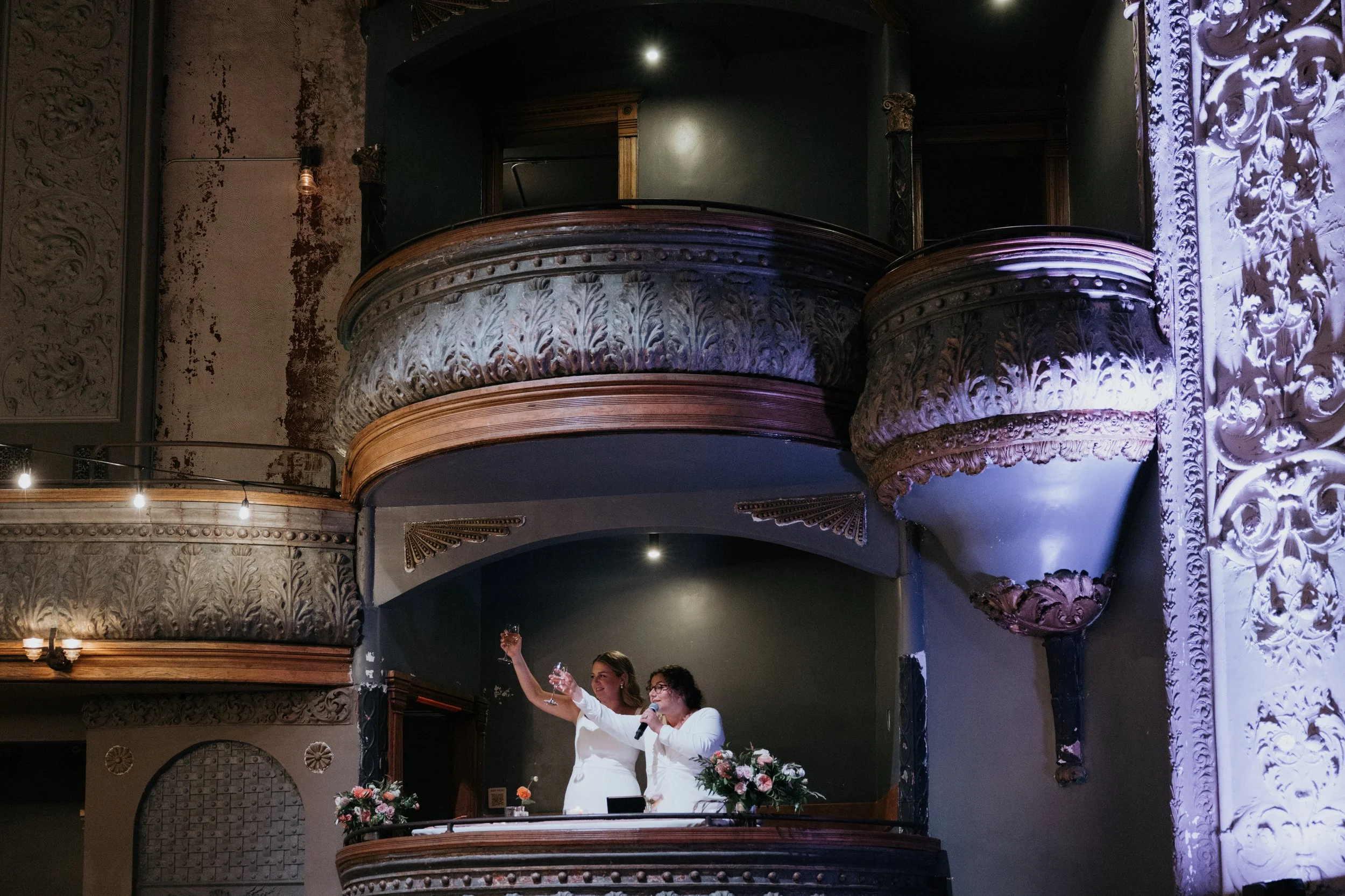 Two women standing on a decorative balcony, one raising a glass, in a room with ornate architecture and floral decorations.