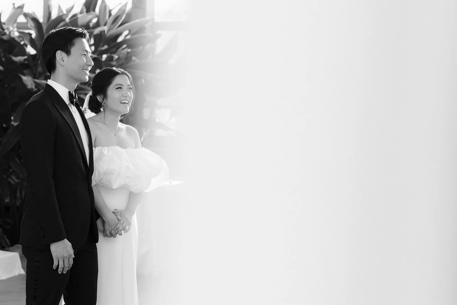 A black and white photo of a bride and groom standing together. The groom is wearing a tuxedo and smiling, the bride is wearing a white dress with ruffled sleeves and smiling.
