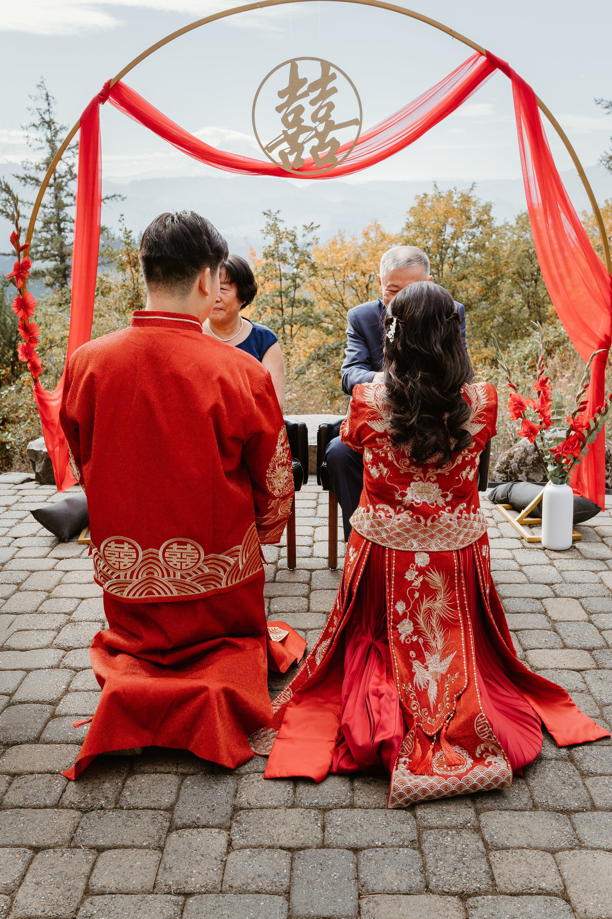 A traditional Chinese wedding ceremony with the bride and groom kneeling on a paved outdoor area, facing each other, with a ceremony officiant and a parent or elder seated behind them. The bride is wearing a red embroidered dress with intricate gold 