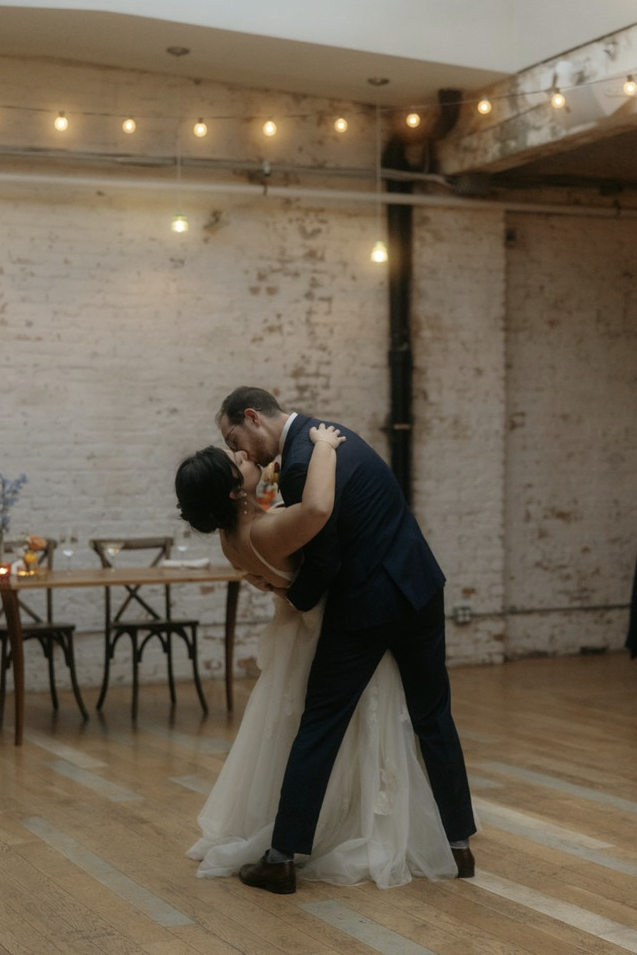 A couple dancing closely in a warmly lit, rustic wedding venue with brick walls and wooden floors.