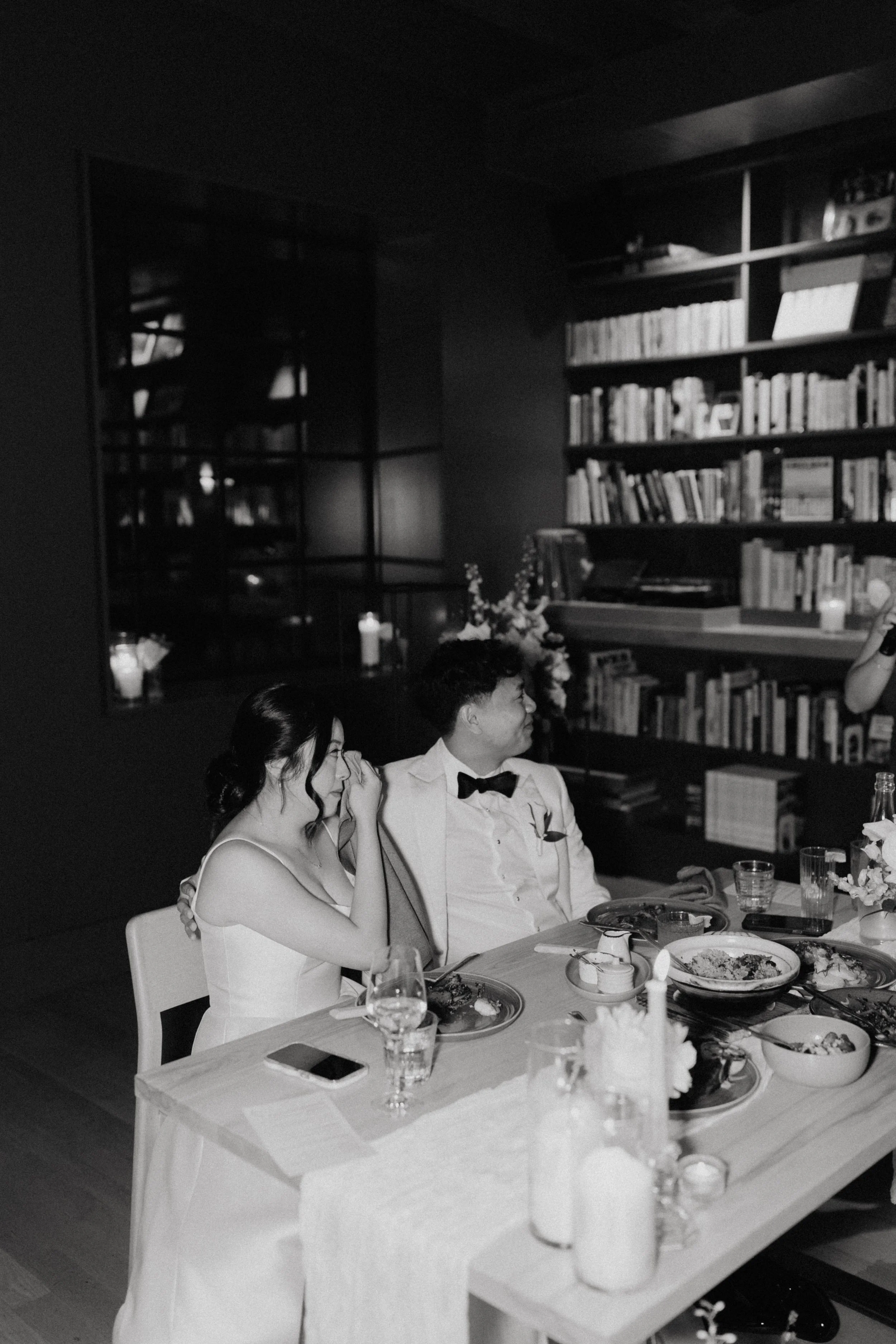 A black and white photo of three people sitting at a table during a dinner, with glasses, plates, and a floral centerpiece. Behind them, there is a bookshelf filled with books and candles on a shelf.