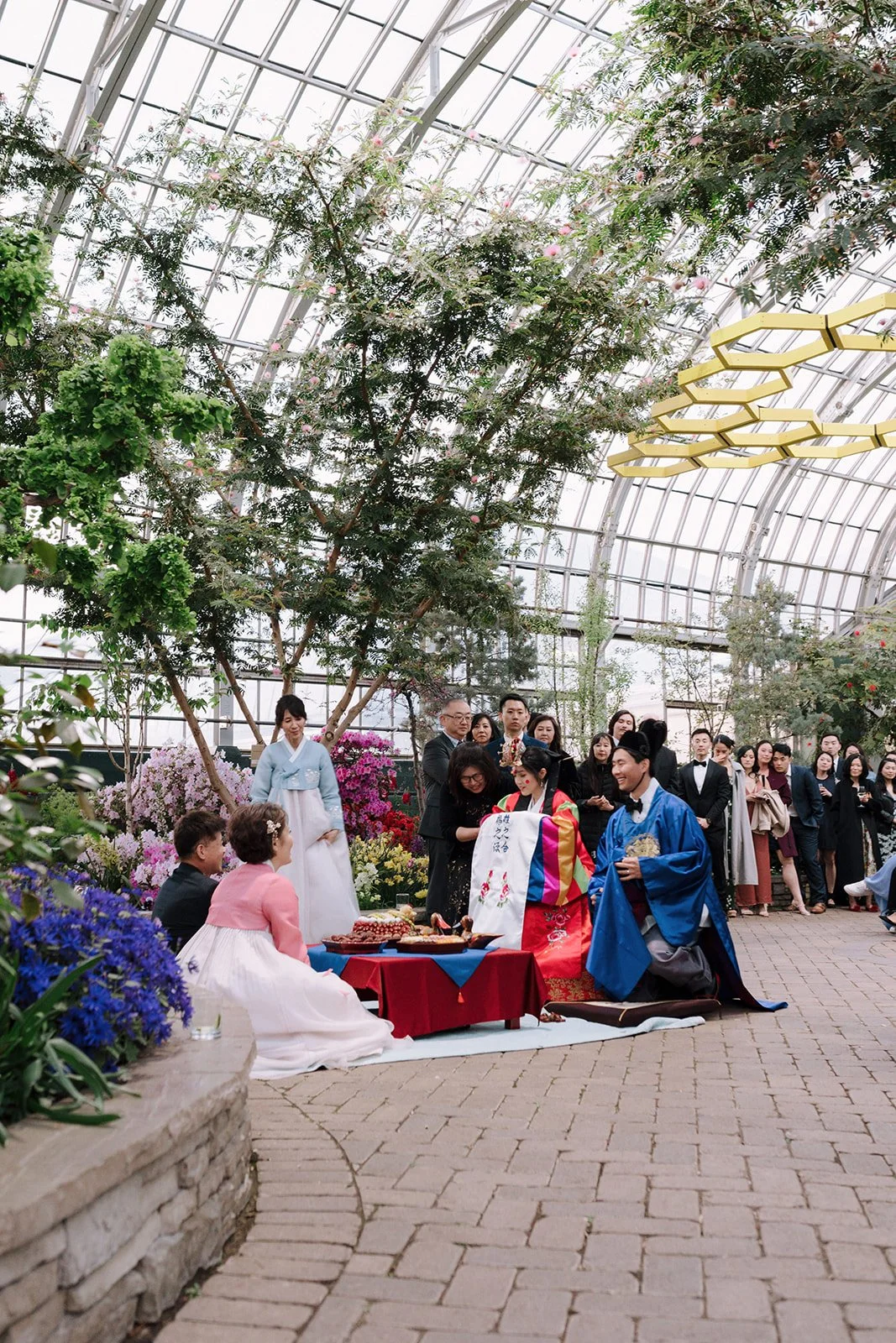 Korean wedding ceremony taking place inside a greenhouse with glass ceiling, surrounded by colorful flowers and trees. The bride and groom are dressed in traditional Korean hanbok, seated on cushions with a ceremonial table in front. Several guests s