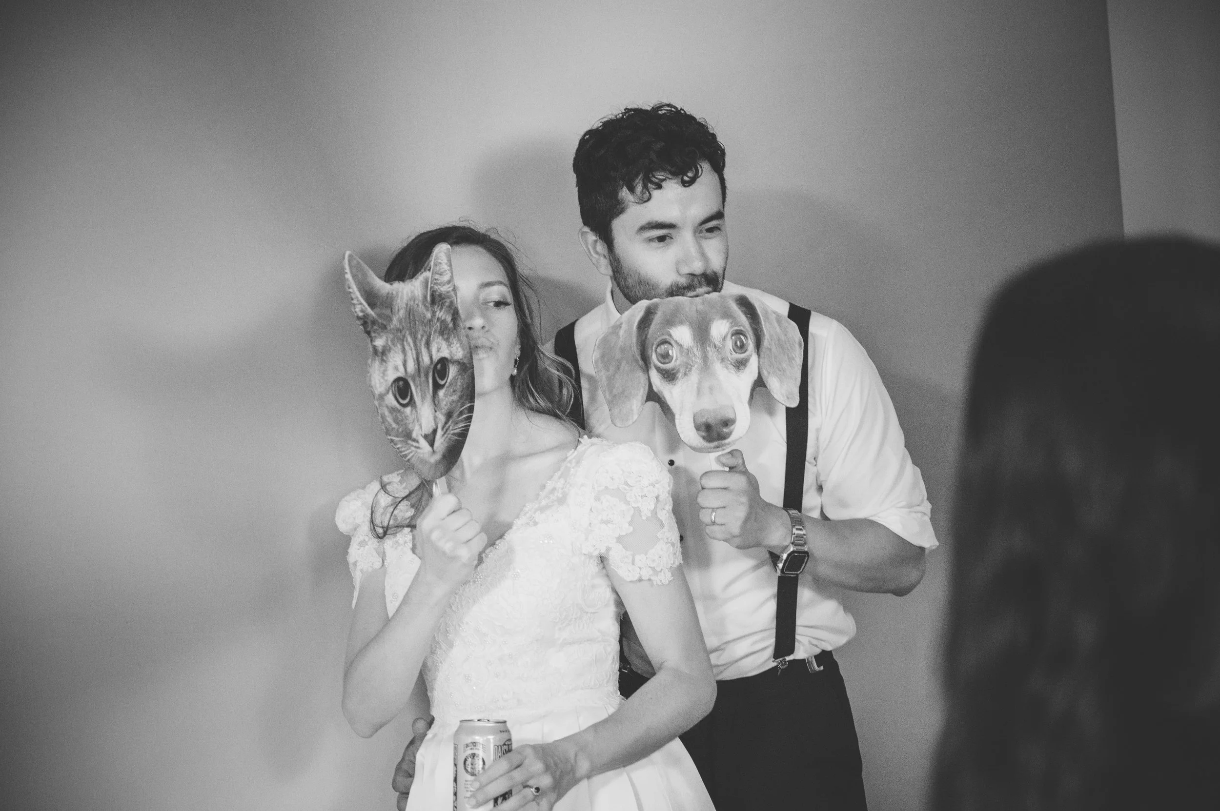 A black and white photo of a couple holding masks of a cat and a dog, with the woman in a wedding dress and the man in a dress shirt and suspenders, standing against a plain wall.