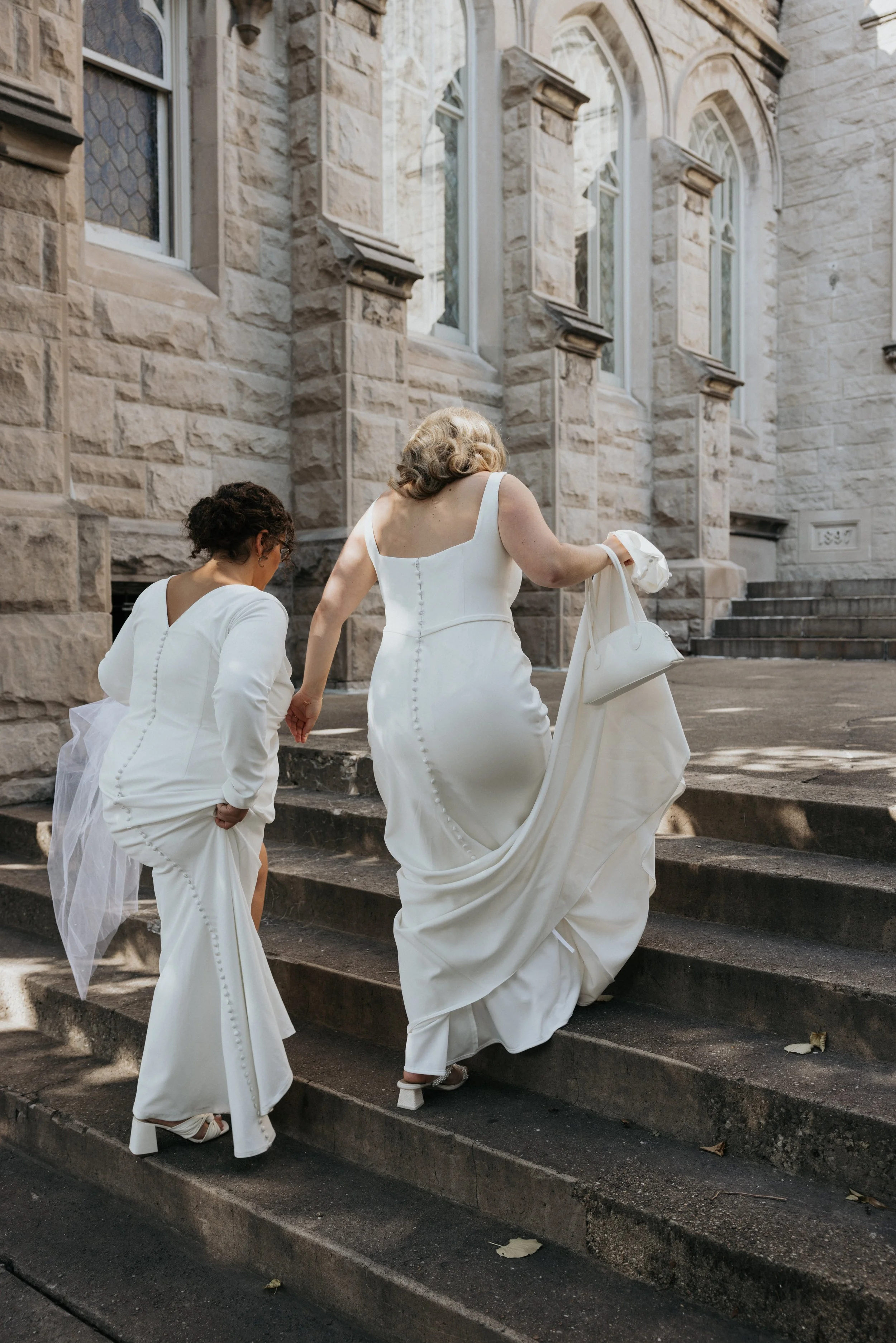 Three women in white dresses walking up stone steps outside a historic stone building with arched windows.