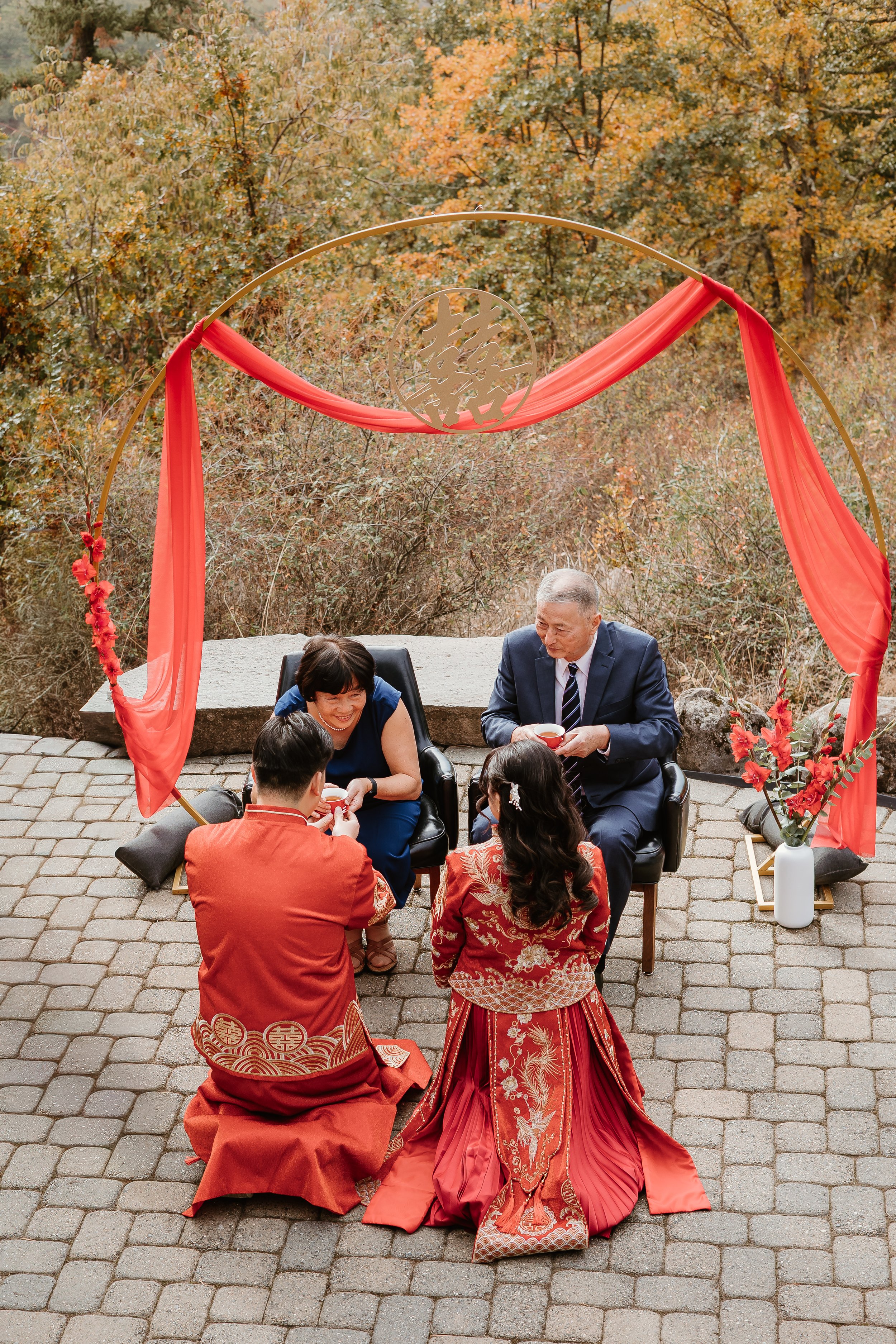 People participating in a traditional Asian wedding ceremony outdoors under a decorative arch with red cloth, surrounded by autumn foliage.