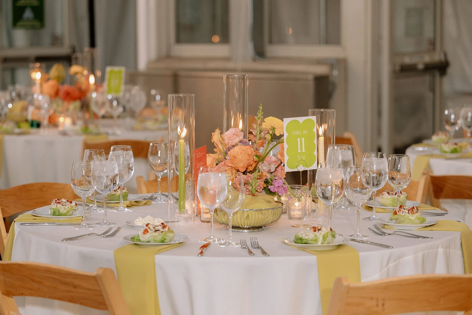 Round banquet table decorated with a floral centerpiece, surrounded by empty water glasses, wine glasses, and plates of salad, set for a formal event or wedding reception in an industrial-style venue.