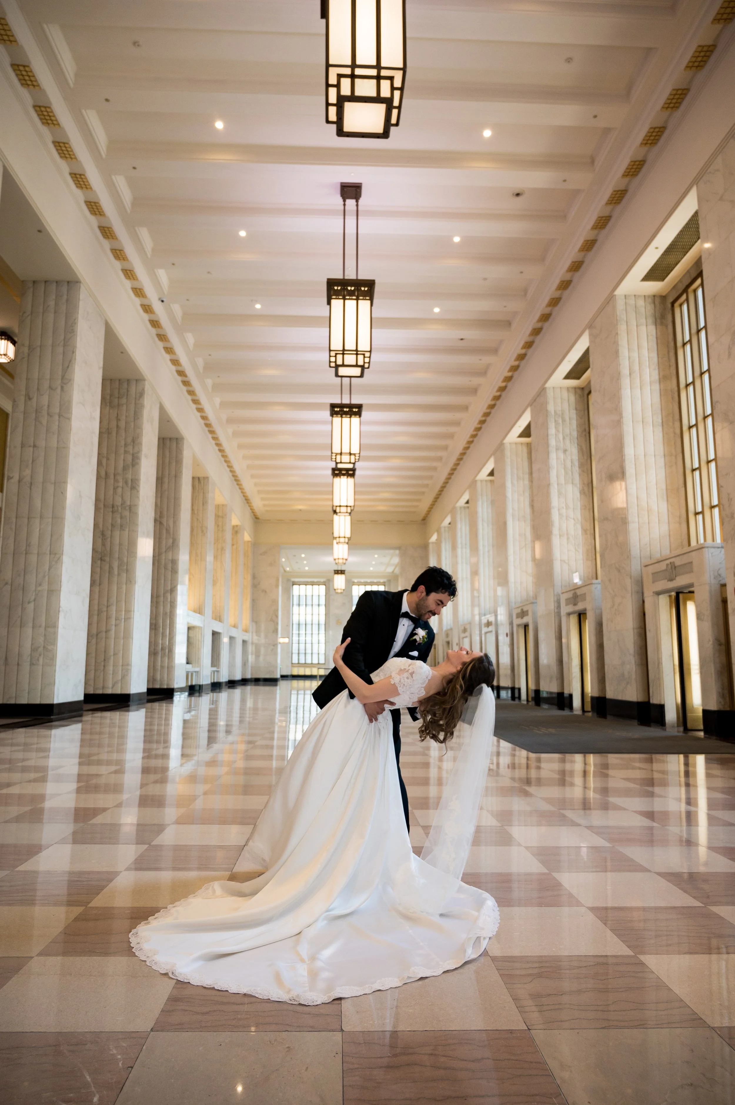 A groom dips and kisses a bride who is leaning back in a grand, well-lit hall with tall columns and hanging pendant lights. The bride is wearing a long white wedding gown with a veil, and the groom is in a tuxedo.
