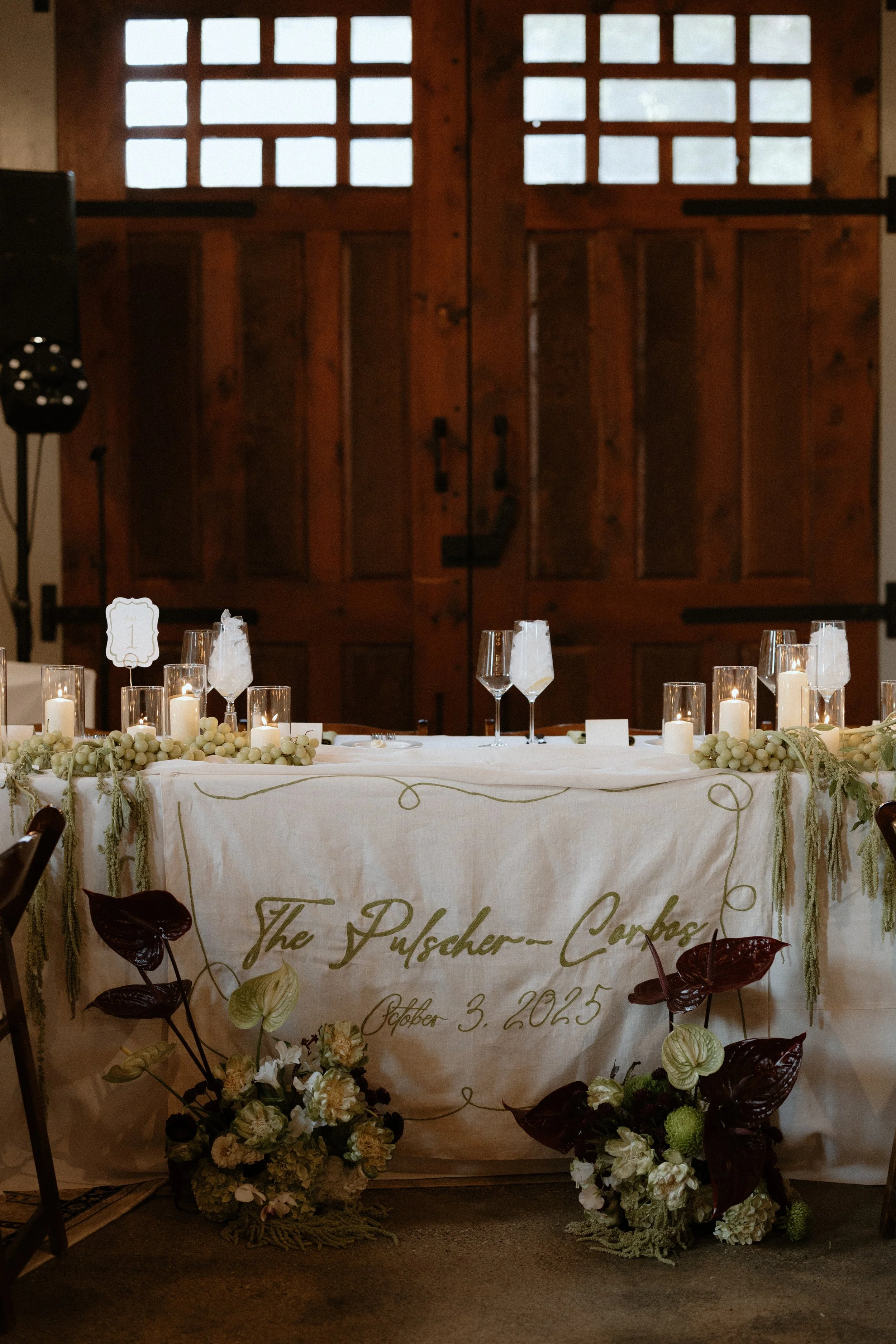 A decorated table with candles and floral arrangements, with a cream-colored cloth and a sign with cursive writing indicating a wedding date, in front of a large wooden barn-style door with windows at the top.