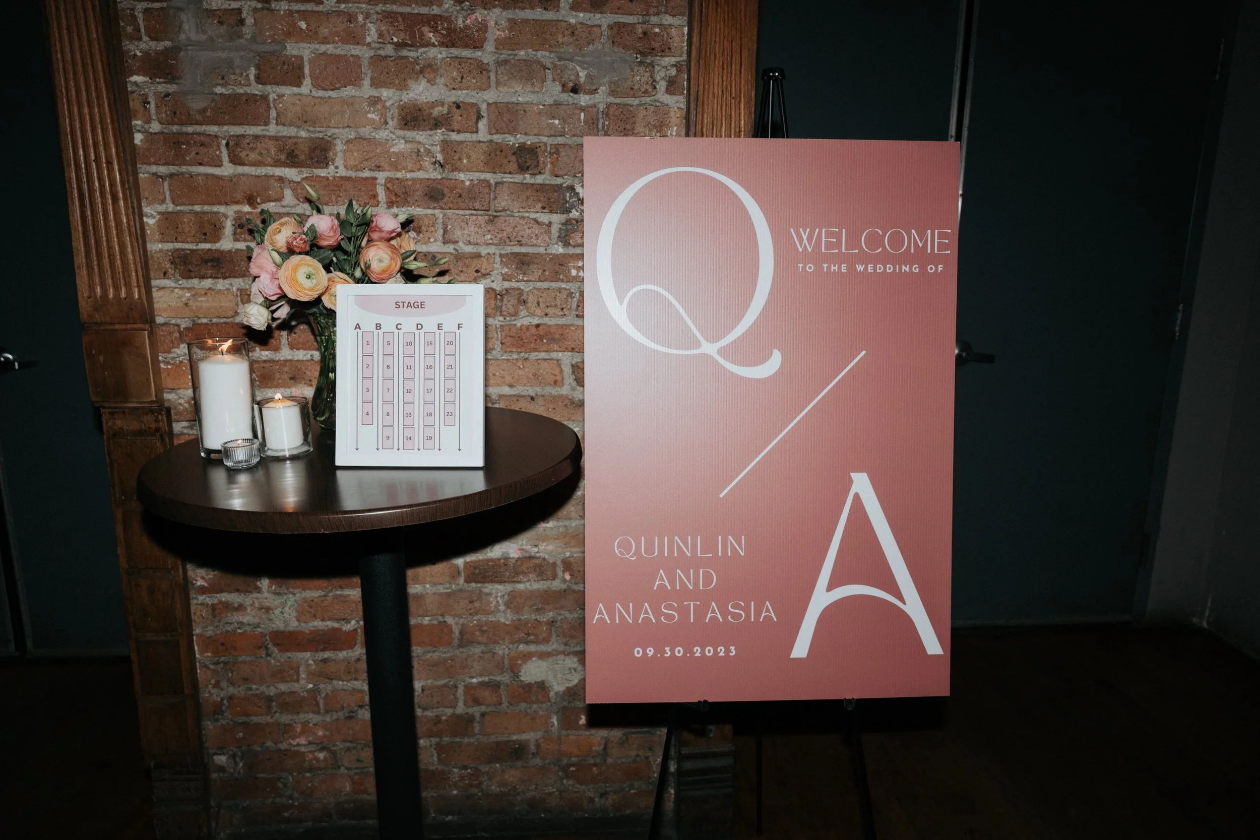 A welcome sign for a wedding event featuring the names Qulin and Anastasia, with the date 09.30.2023. The sign has a pink and white color scheme. Nearby is a small round table with a flower arrangement of pink and cream flowers, three candles, and a 