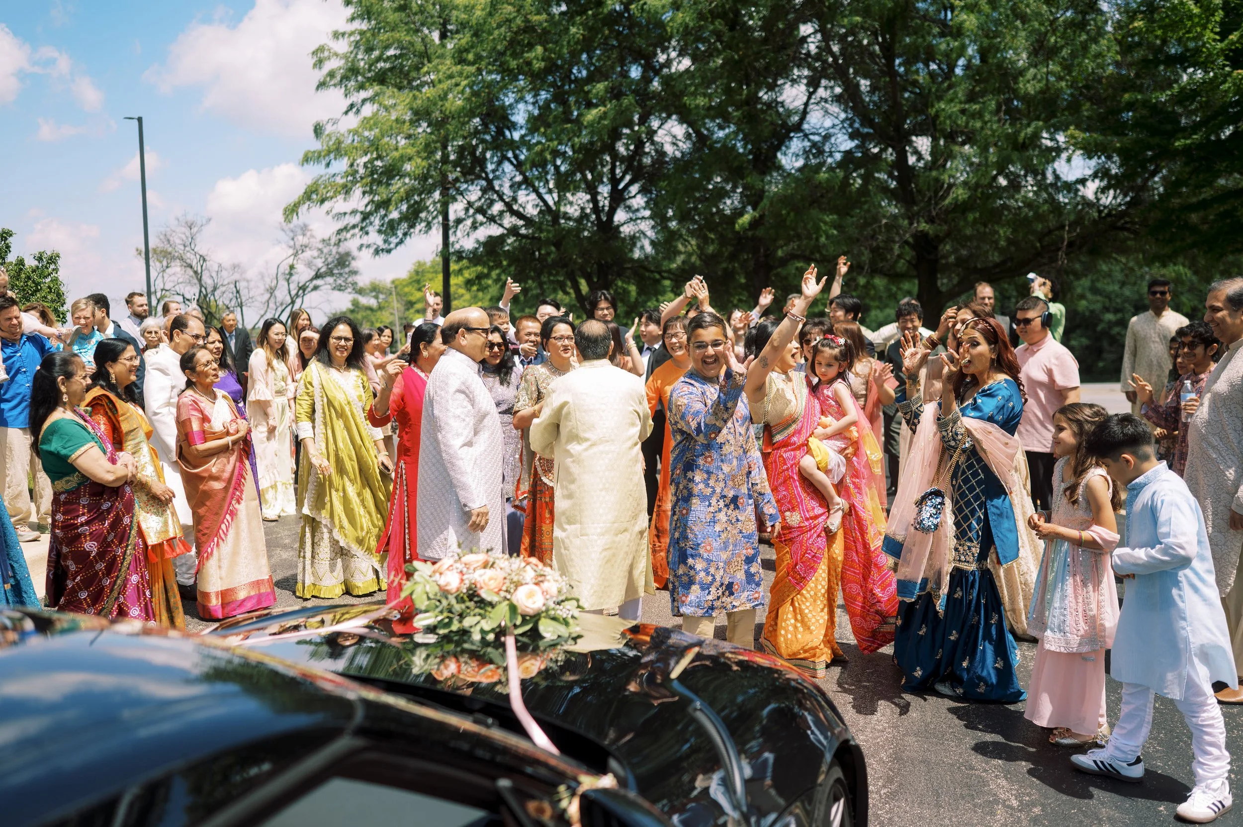 A large group of people dressed in colorful traditional South Asian clothes gathered outdoors, celebrating with smiling faces and raising their hands, with lush green trees and cloudy sky in the background.