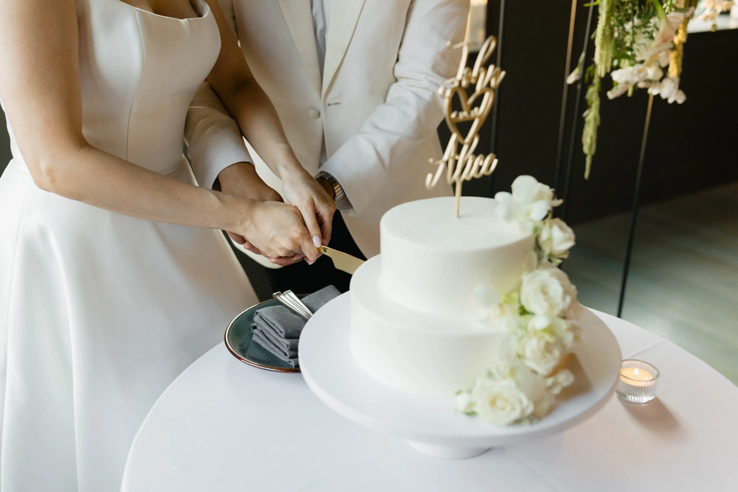 Close-up of a couple cutting a wedding cake at a celebration. The cake is decorated with white flowers and a 'love' cake topper. The bride is wearing a white dress, and the groom is in a white suit.