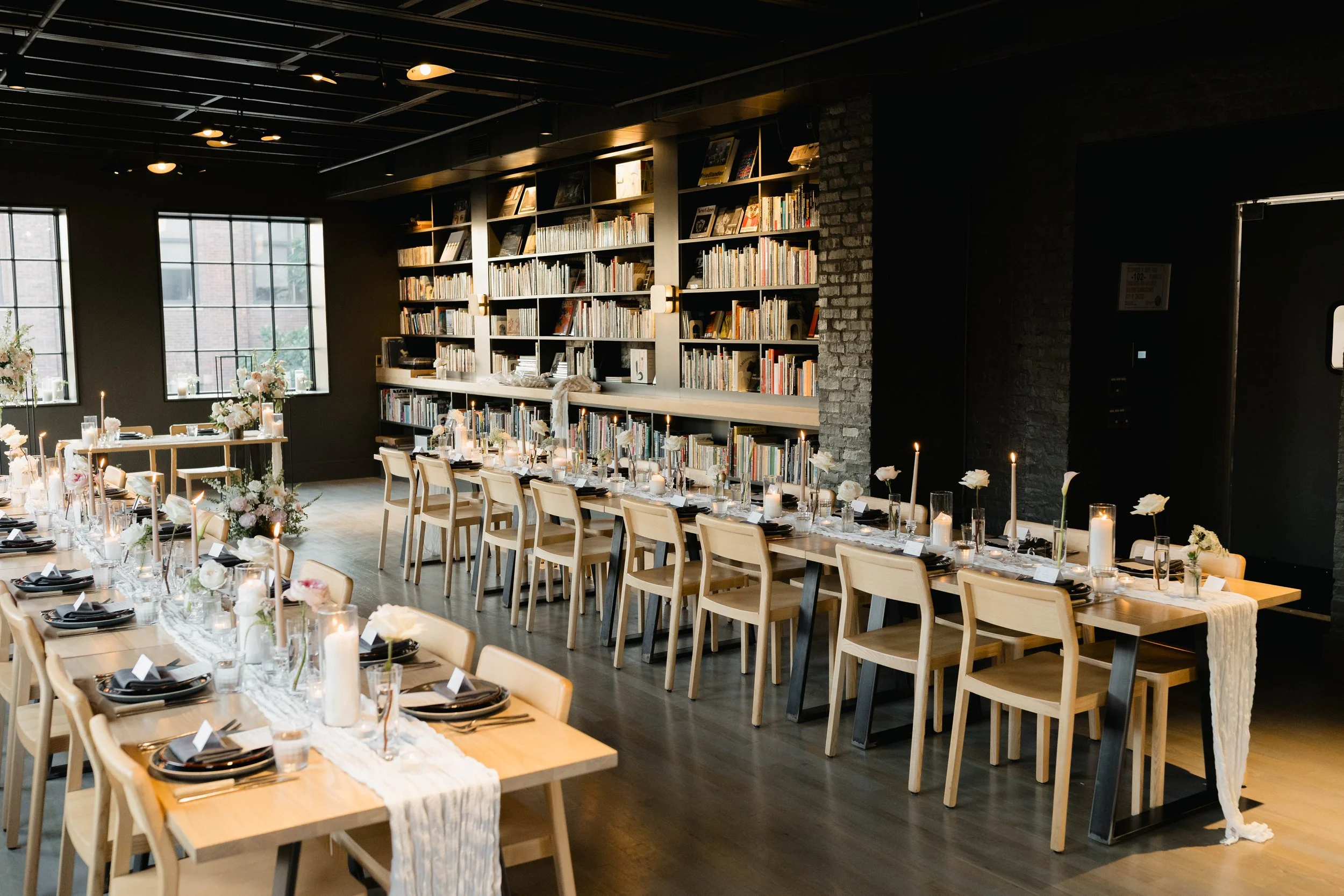 A dining area set up for a celebration with tables decorated with flowers and candles, surrounded by bookshelves filled with books in a dimly lit room with large windows.
