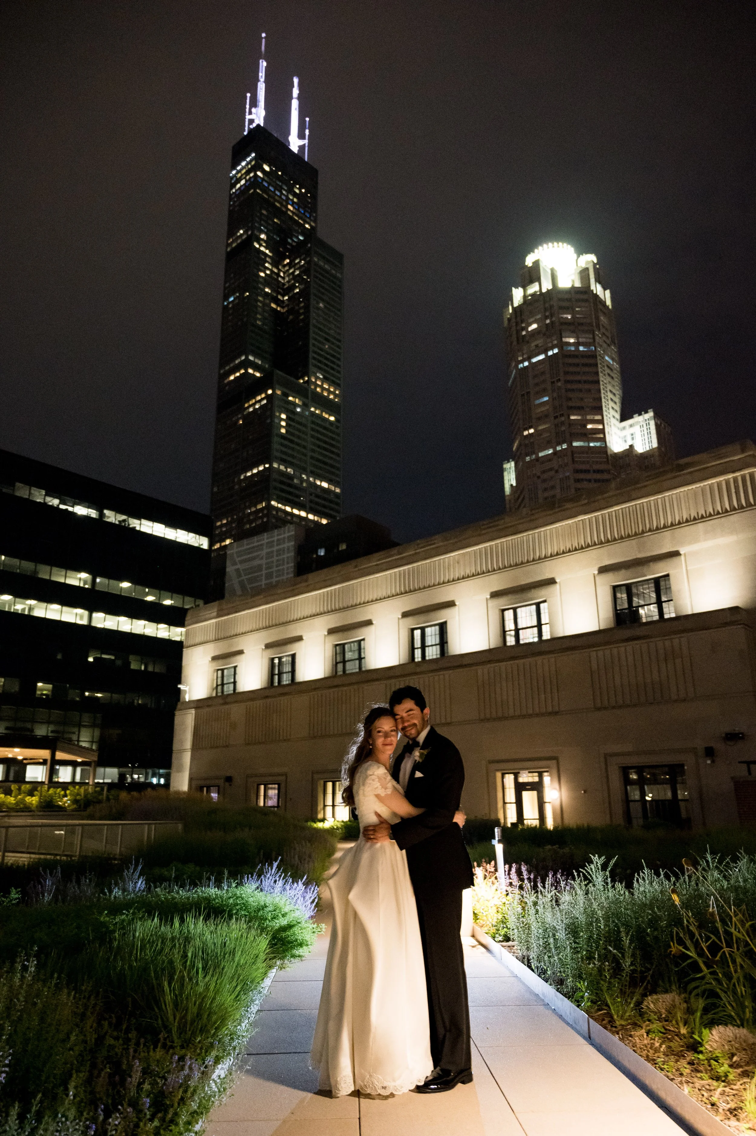 Happy couple in wedding attire embracing on a city rooftop at night, with illuminated skyscrapers in the background.
