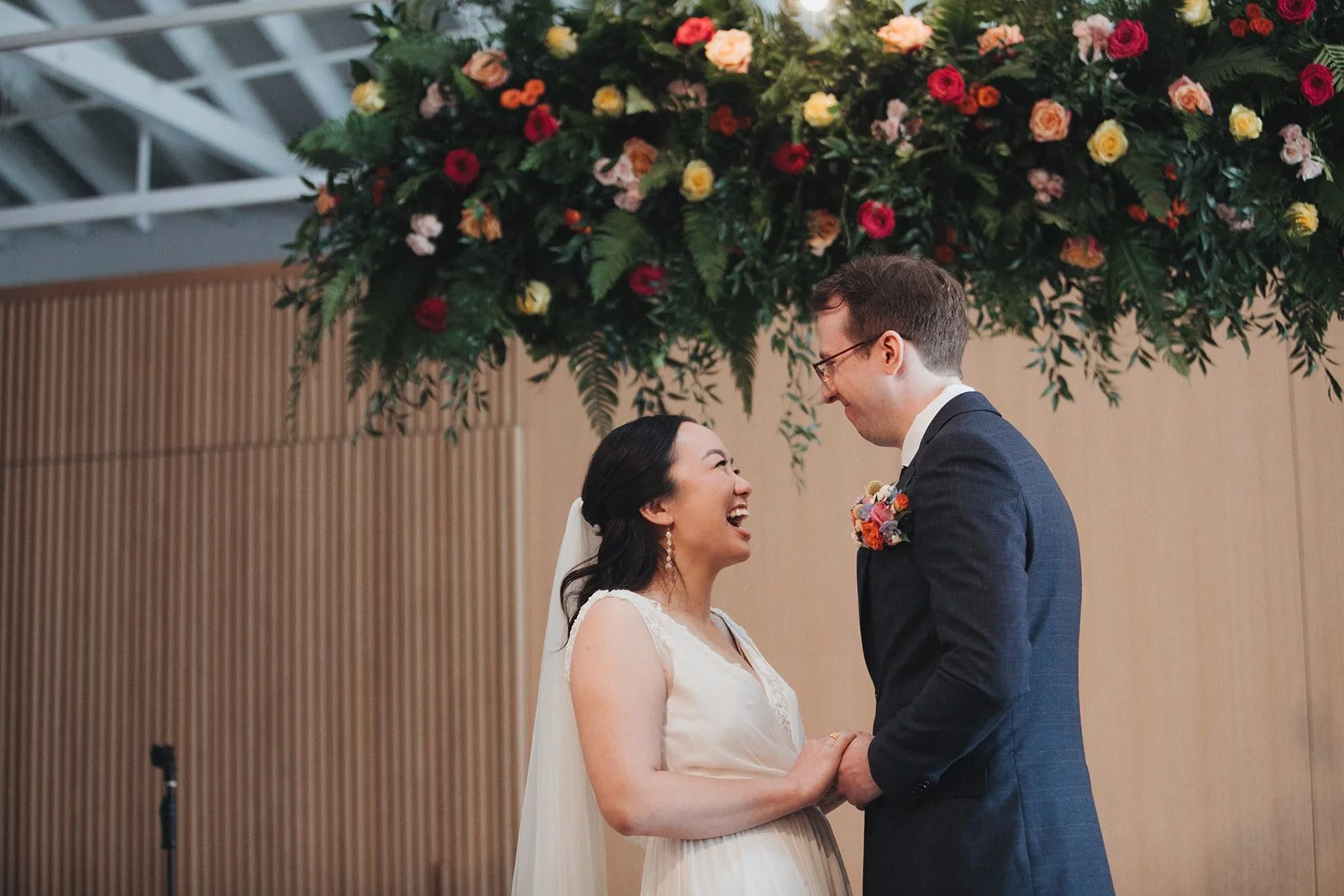 A bride and groom holding hands and smiling at each other during their wedding ceremony under a large floral arrangement with red, pink, and yellow flowers.
