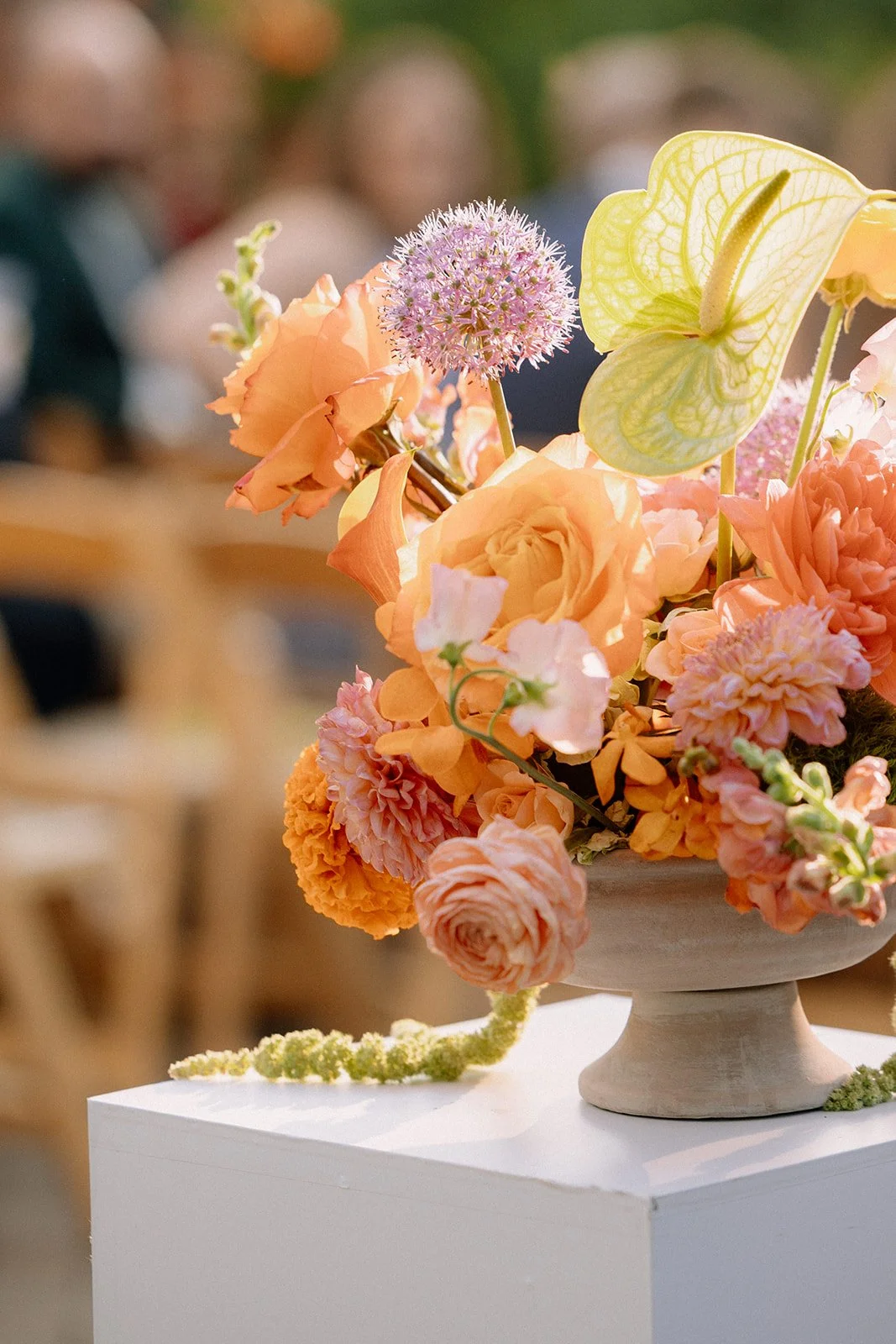 A floral arrangement in a beige vase with peach, pink, and green flowers, including a yellow bell-shaped flower and a purple spherical flower, on a white pedestal.