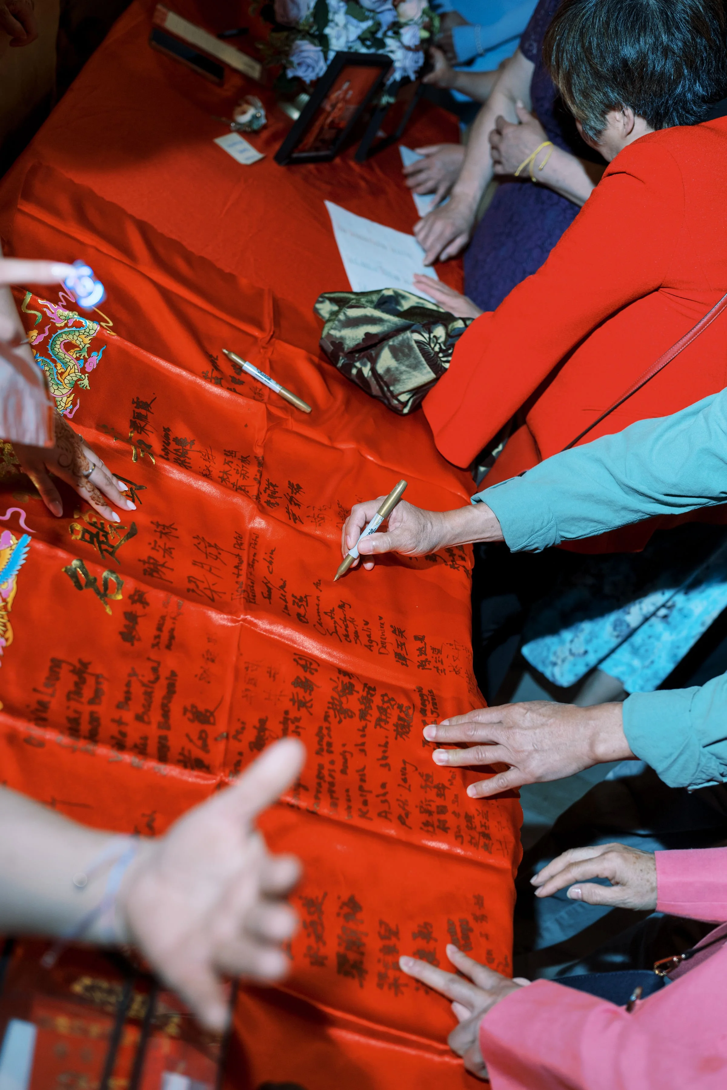 People signing a red banner with Chinese characters, some holding pens, in a celebratory or commemorative event.