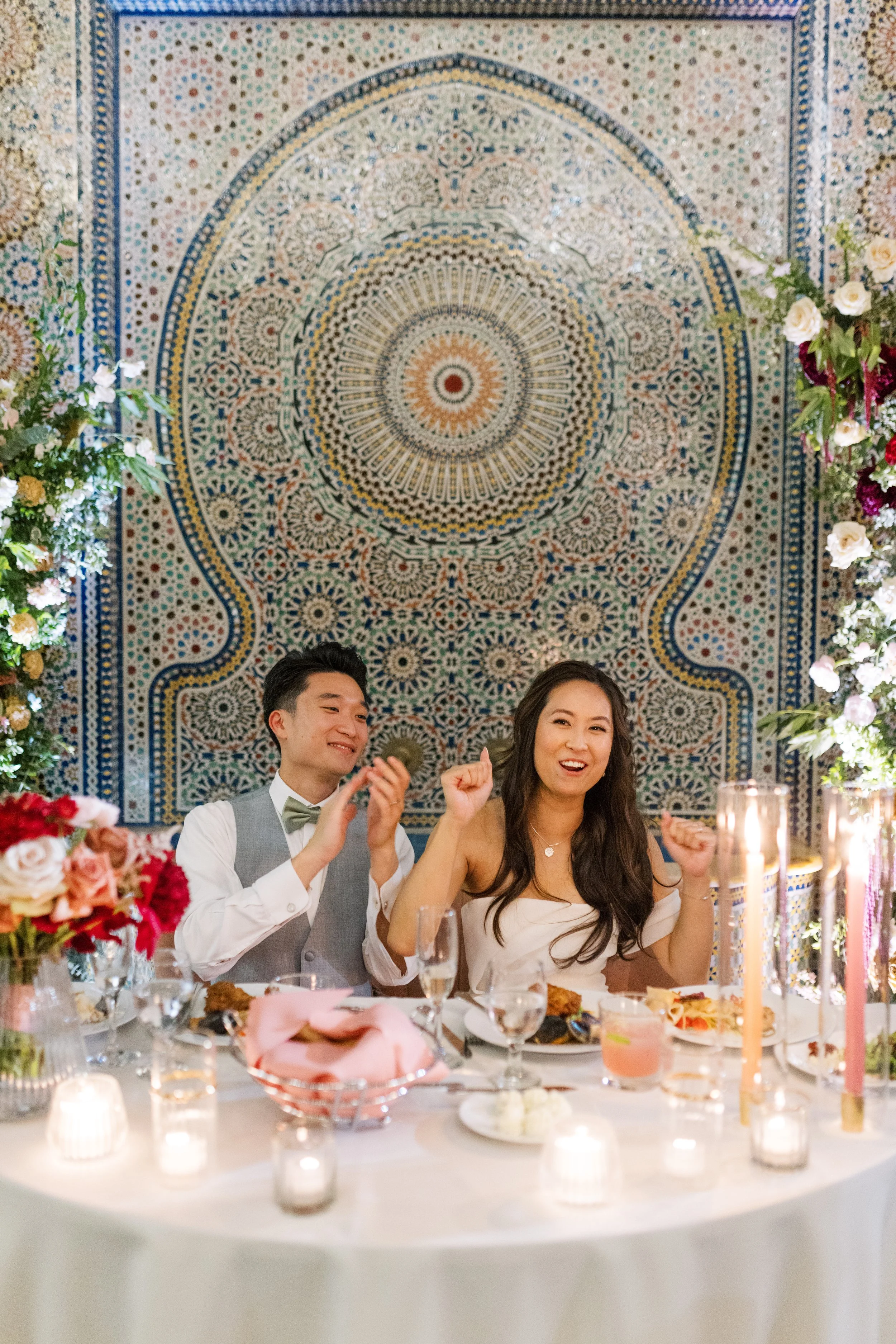 A joyful couple celebrating at a decorated wedding reception table with floral arrangements, candles, and food, set against a colorful mosaic wall.