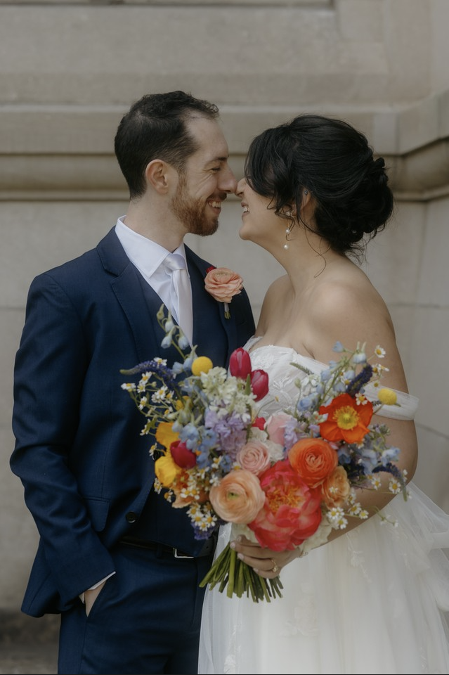 A happy couple at their wedding with their foreheads touching, smiling, and holding a vibrant bouquet of flowers in front of a stone building.