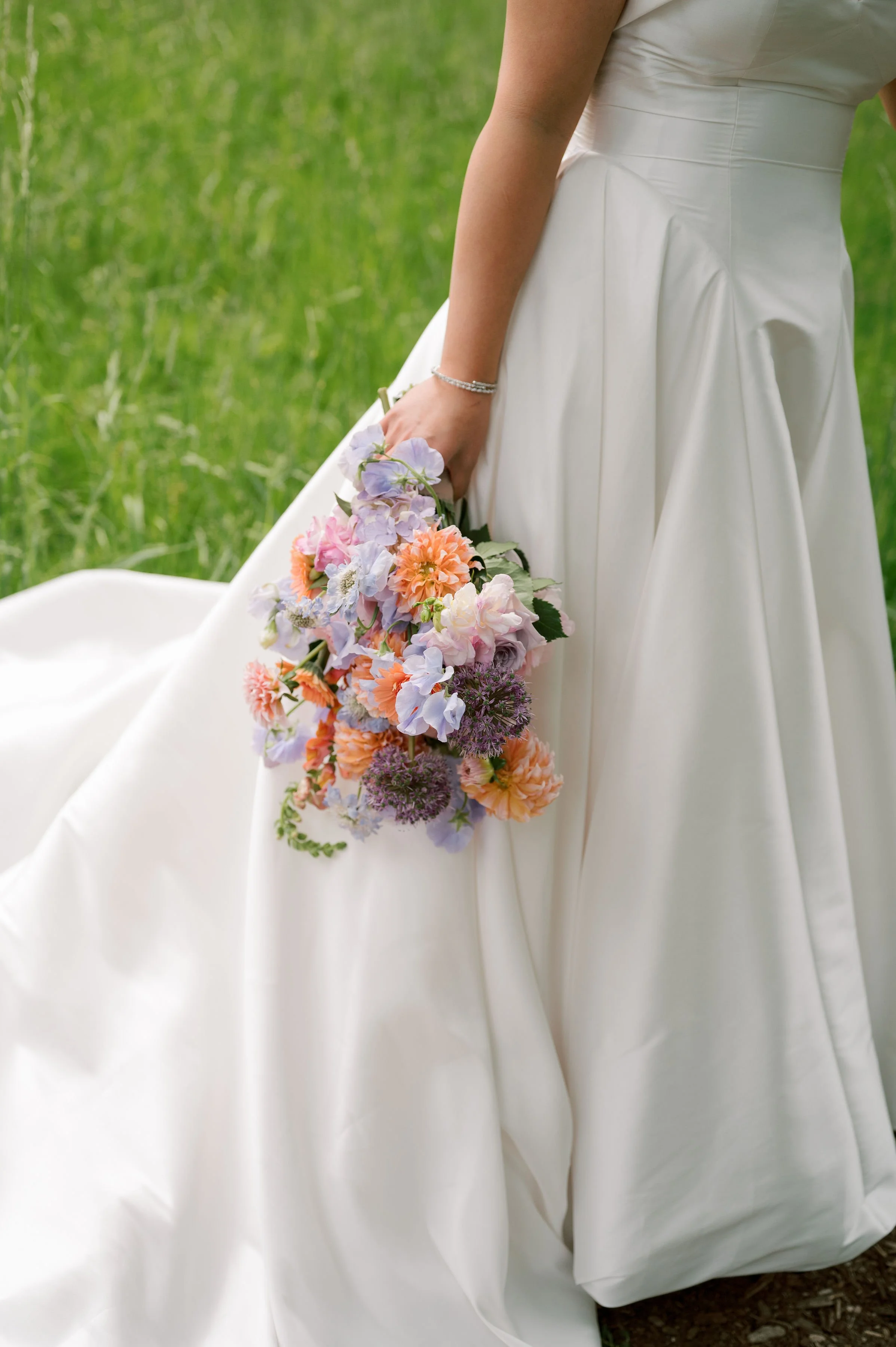 Close-up of a bride holding a bouquet of colorful flowers, wearing a white wedding dress, outdoor setting with green grass.
