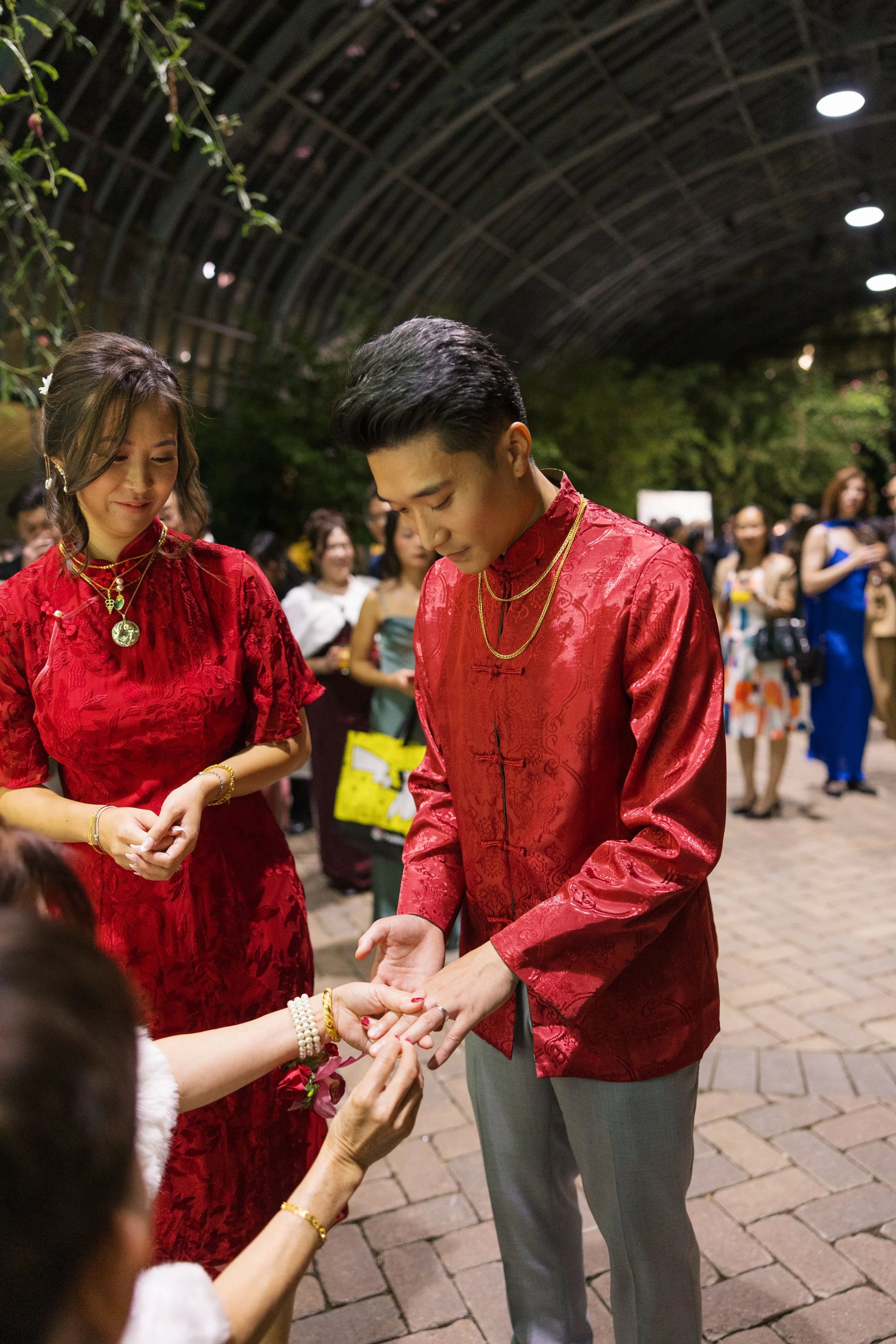 A couple dressed in traditional Chinese red attire participating in a wedding ceremony, with a woman presenting rings and guests in the background under a large arched structure.