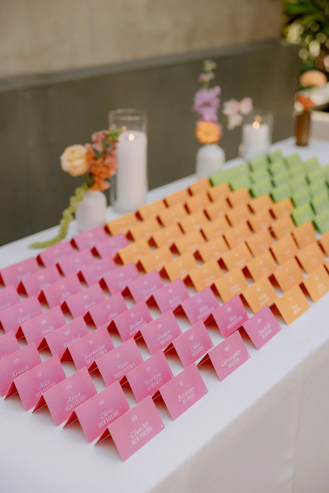 Colorful folded place cards arranged in rows on a table, with pink, orange, and green cards and floral decorations in glass candle holders in the background.