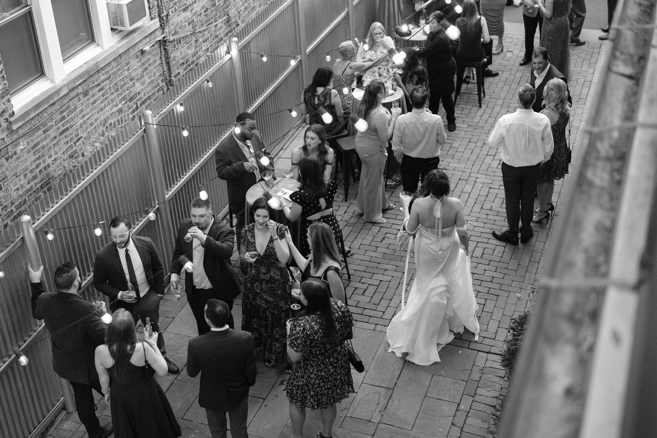 Black and white photo of people socializing outdoors on a brick patio, some holding drinks, illuminated by string lights, with a brick building wall on one side.