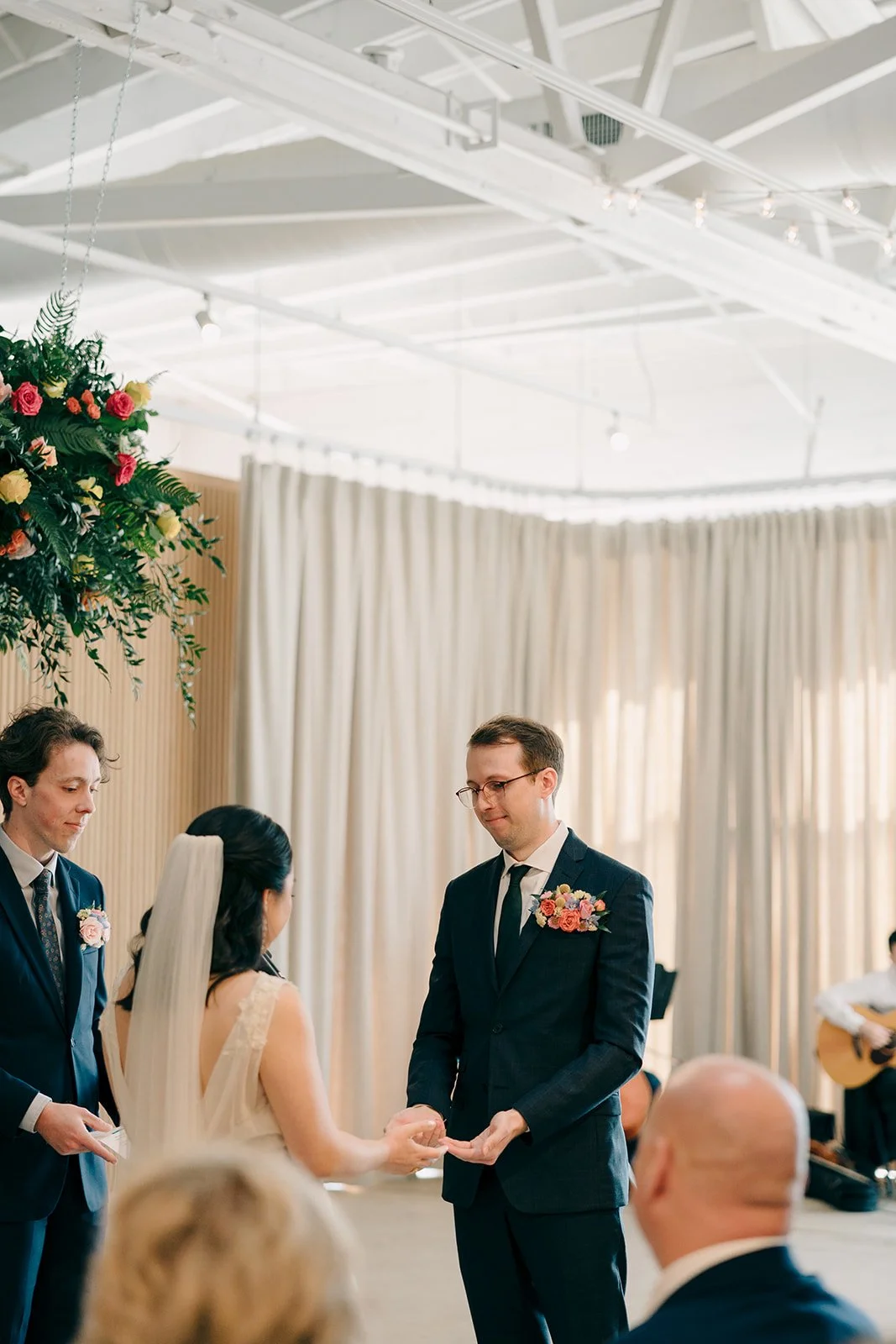 A couple getting married during a wedding ceremony, with the bride and groom holding hands in front of officiant, in a bright indoor venue with beige curtains, floral arrangements, and string lights.