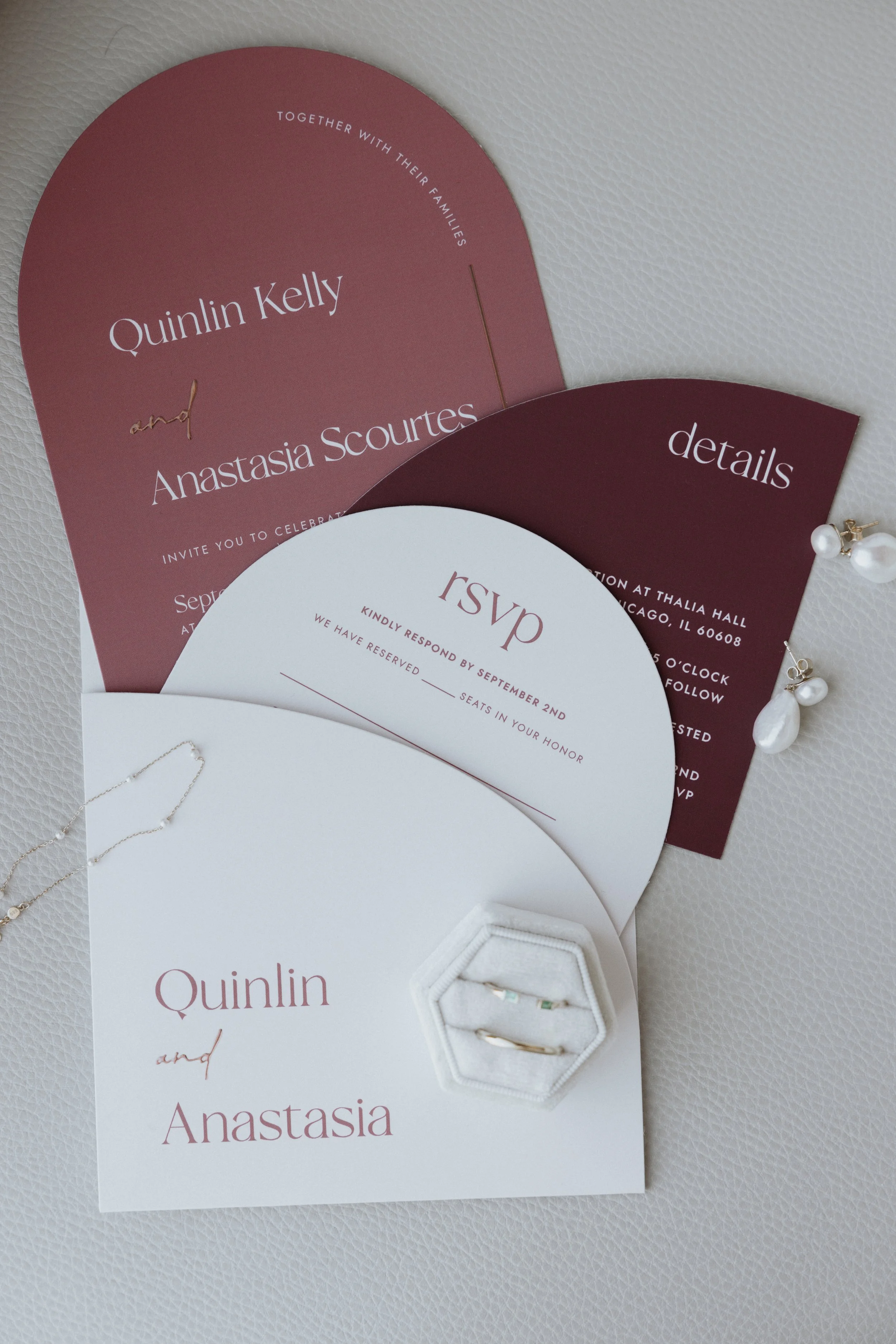 Wedding invitations labeled Quintin Kelly and Anastasia Scourtis, with a pearl jewelry piece and a white jewelry box nearby, on a textured white background.