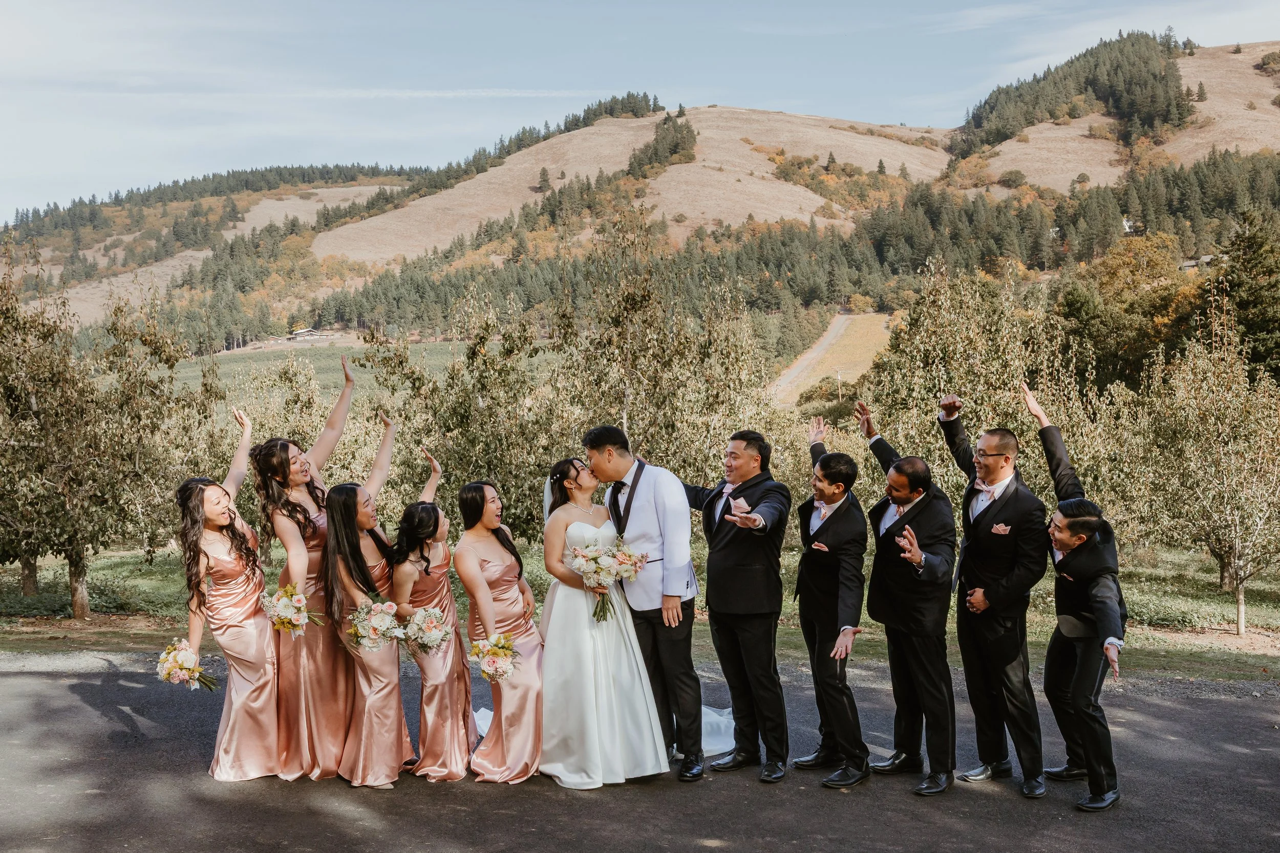 A group of wedding guests celebrating outdoors in a vineyard with hills in the background. The bride and groom are kissing, surrounded by bridesmaids in matching dresses holding bouquets and groomsmen in suits.
