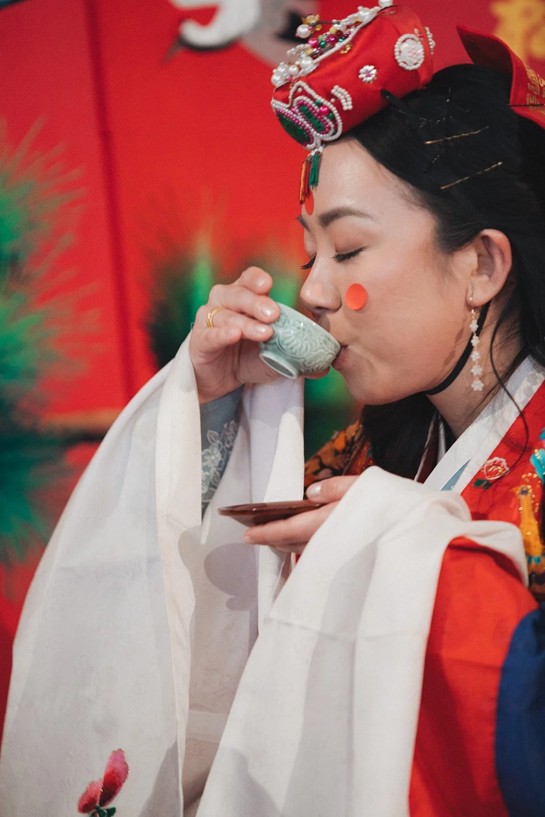 A woman dressed in traditional Korean attire, wearing a colorful hanbok and ornate headdress, drinking tea from a small ceramic cup.