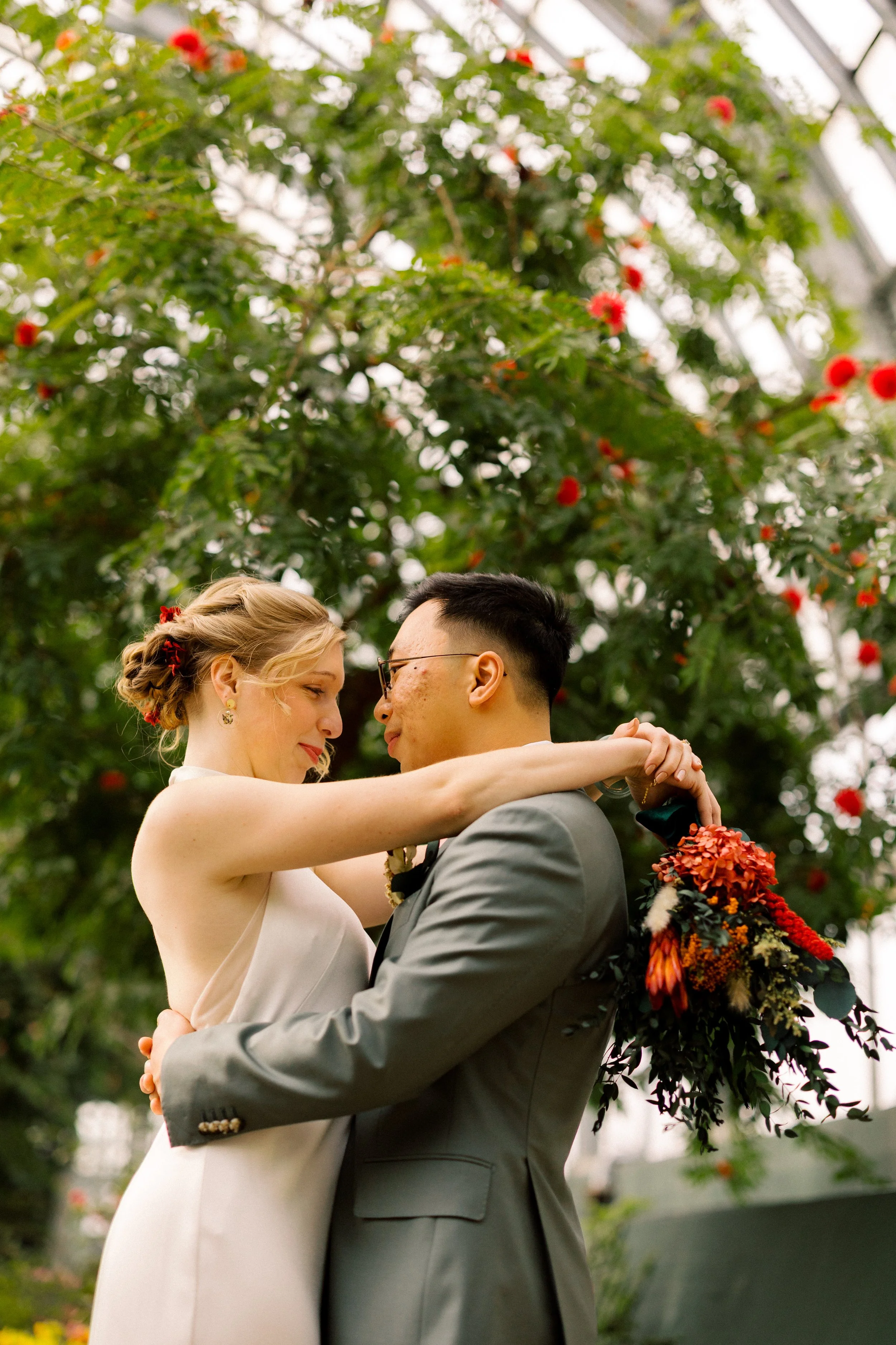 A couple sharing a dance in a greenhouse with lush greenery and red flowers in the background, dressed in wedding attire.