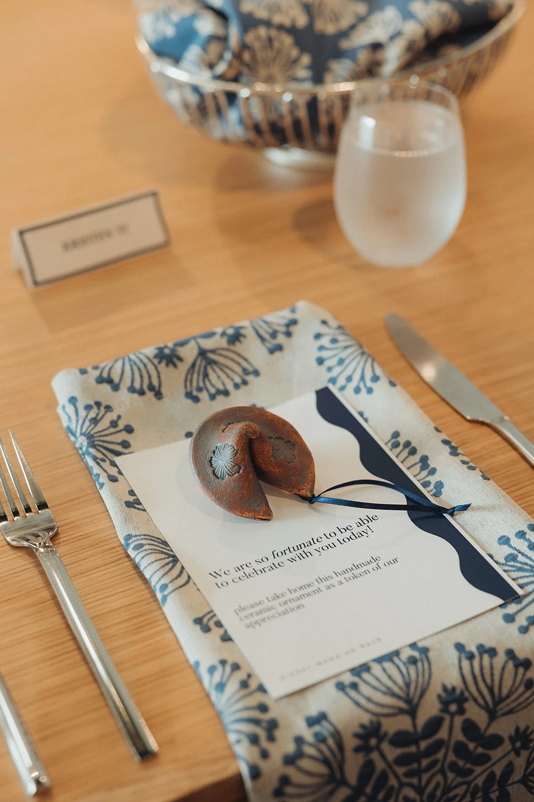 A table setting with a fork on the left, a knife on the right, a decorative linen napkin, a colonial ornament, a card with a message, a glass of water, and a large bowl with a blue and white floral pattern in the background.