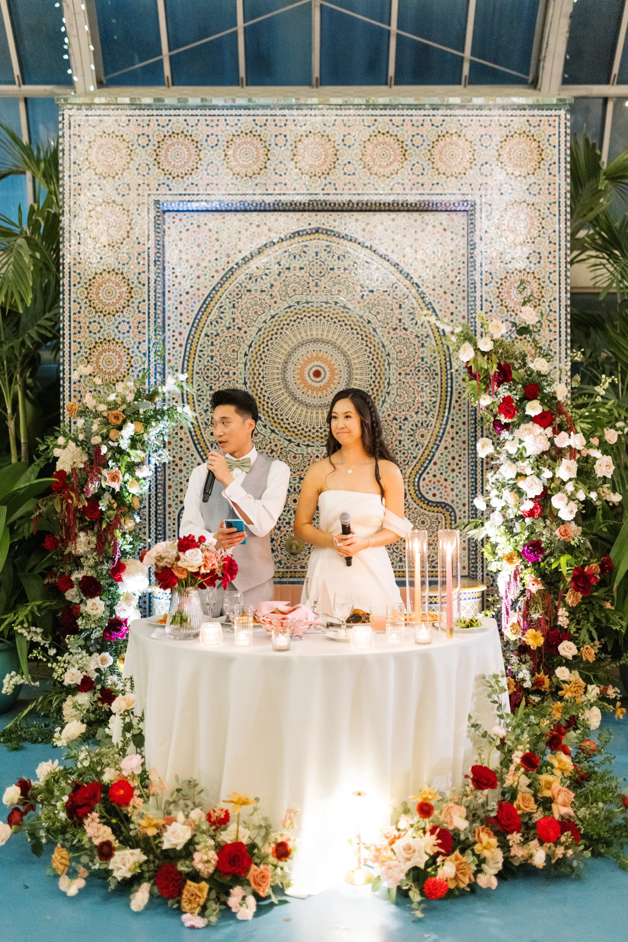 A couple at a wedding reception standing behind a table decorated with flowers and candles, with a large ornate mosaic backdrop.