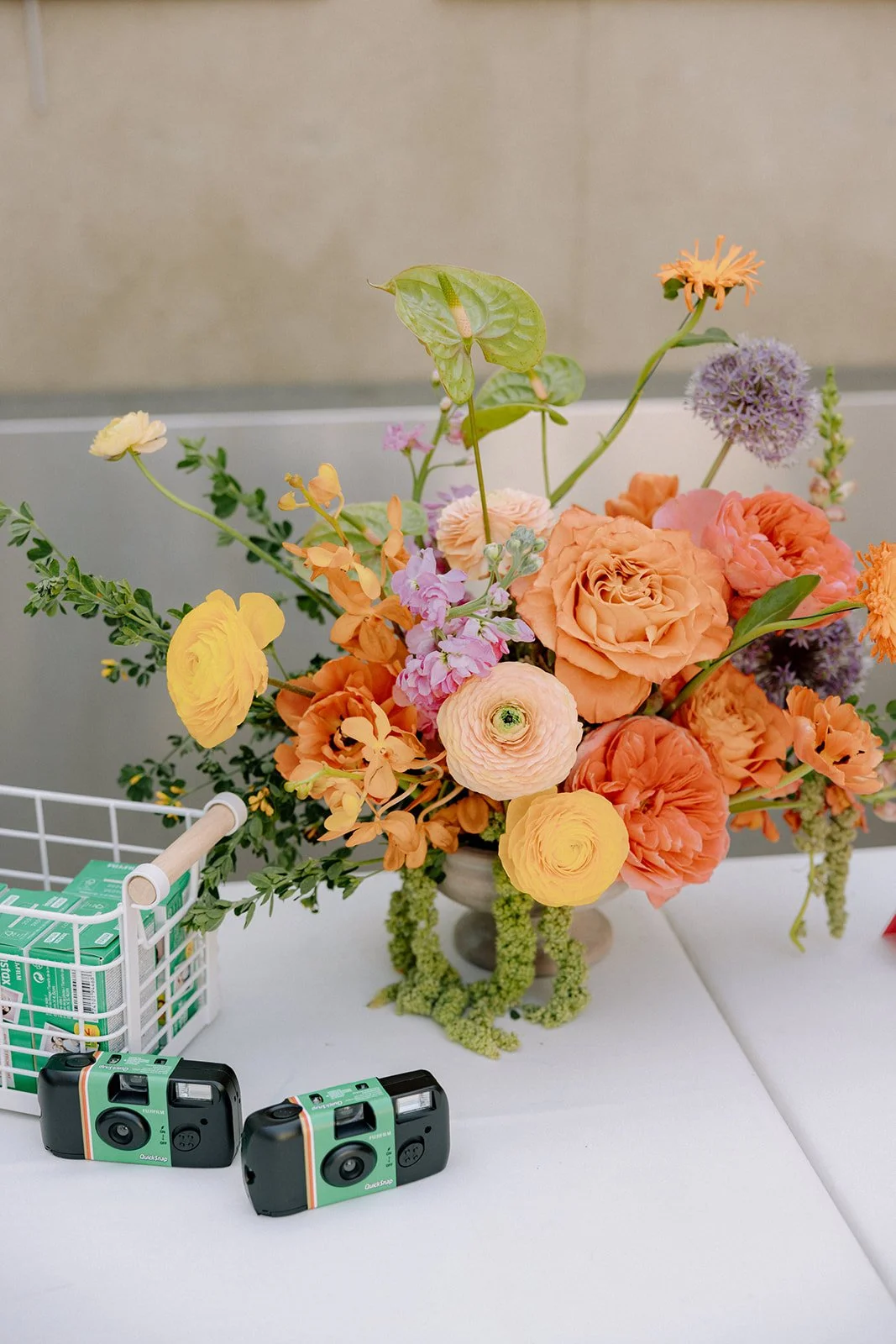 Colorful flower arrangement in a vase, surrounded by vintage cameras and a small white basket with green boxes, on a white table against a beige wall.